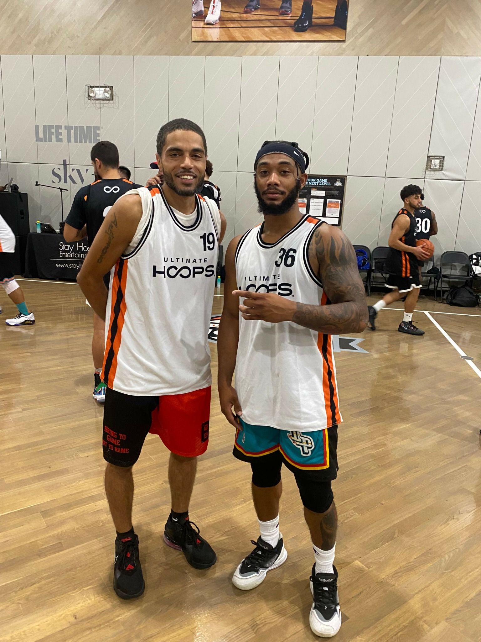 Two basketball players in white jerseys with black and orange accents stand on a wooden court, one giving a thumbs-up, the other smiling.