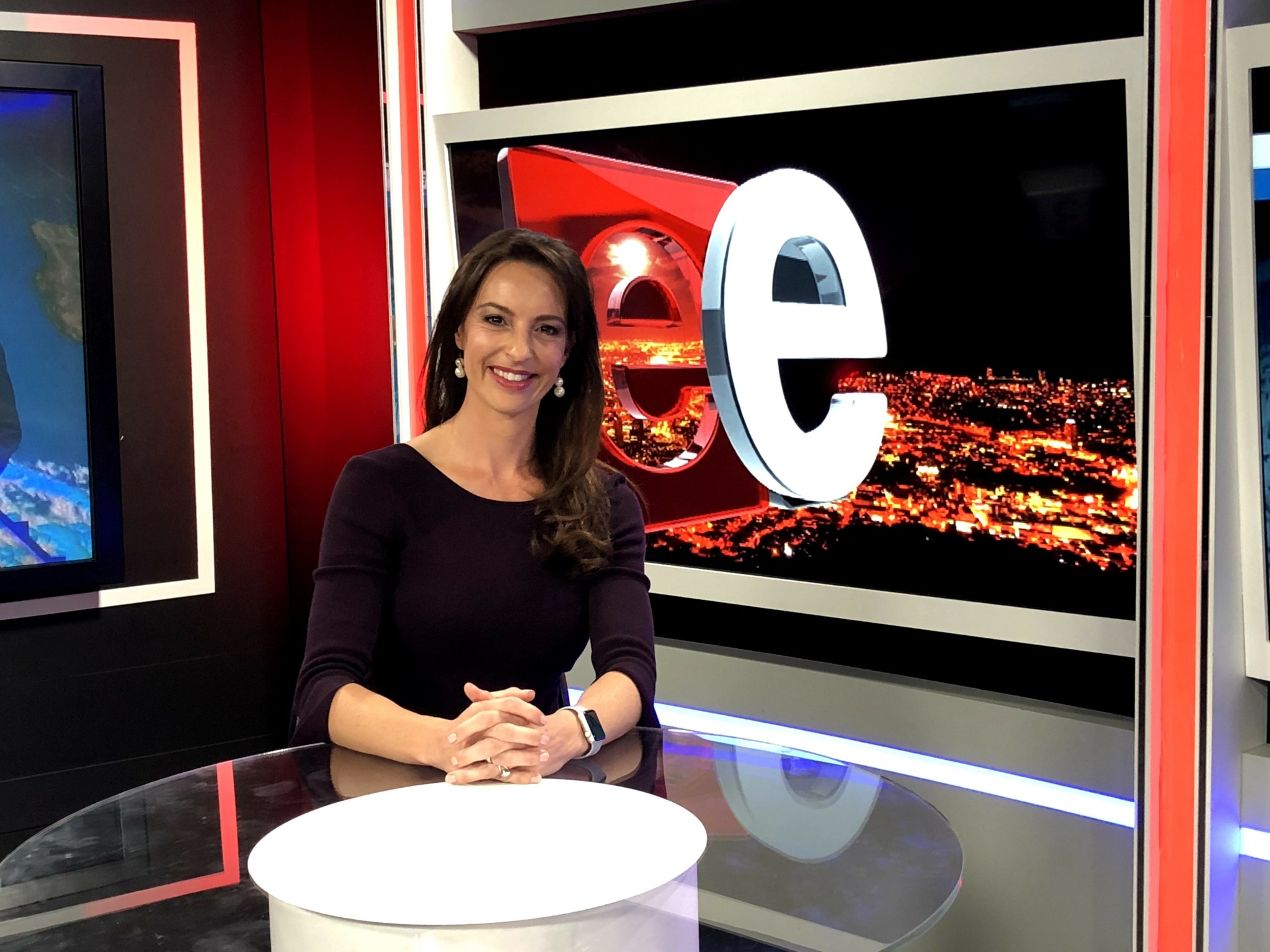 Woman sitting at a news desk with a large illuminated 'e' behind her, city lights visible in the background.