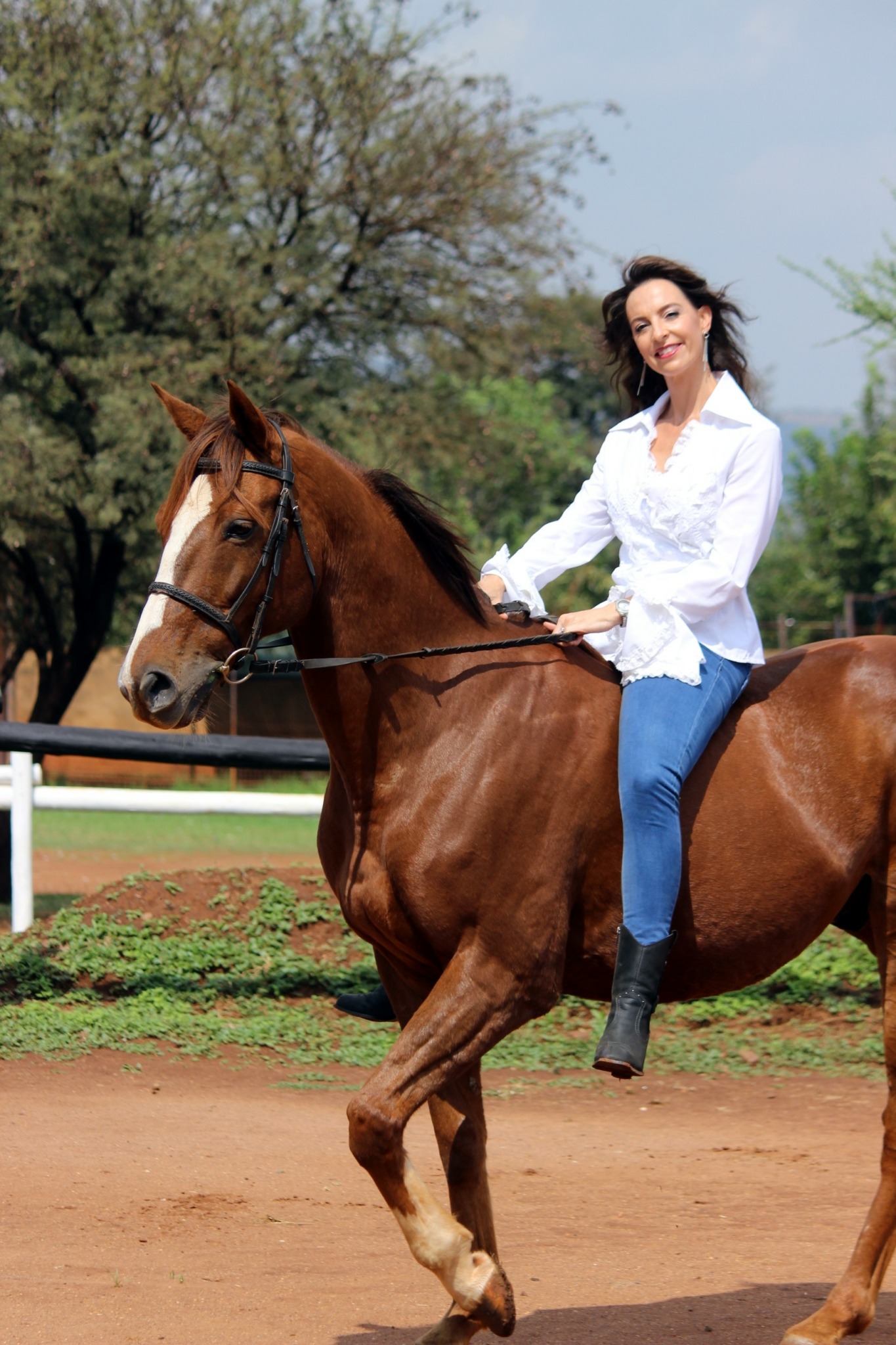 Woman riding a brown horse on a dirt track outdoors, smiling, with trees and a fence in the background.