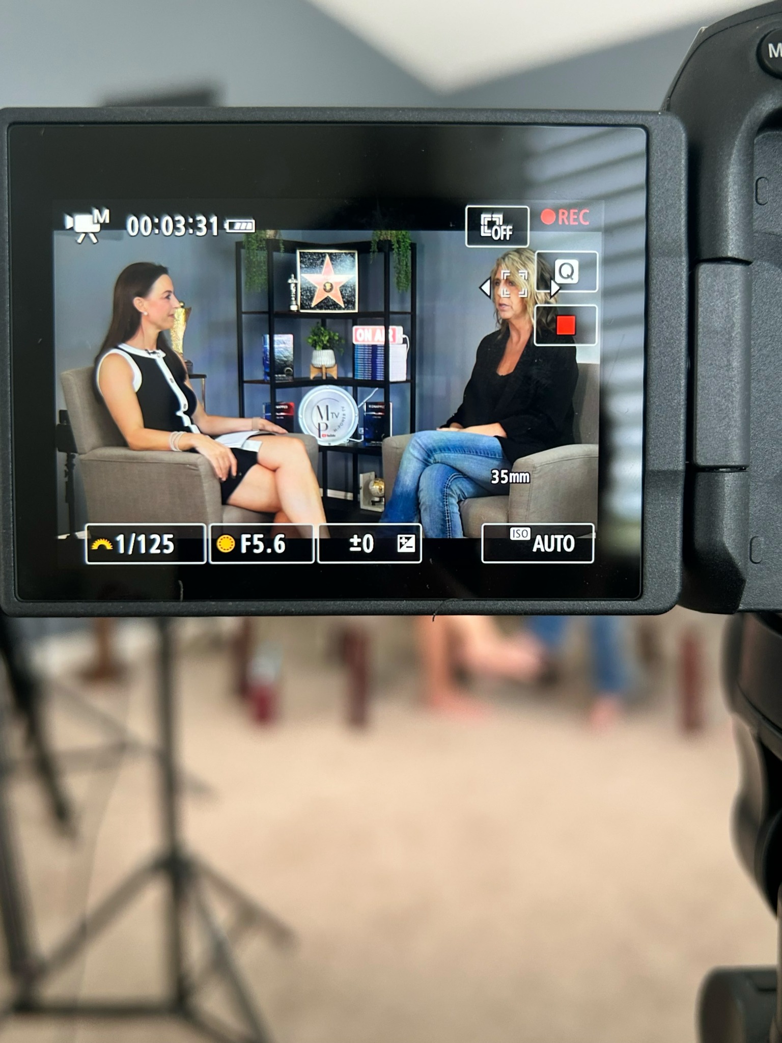 Two women seated and talking in a studio setting with shelves and decorations behind them.