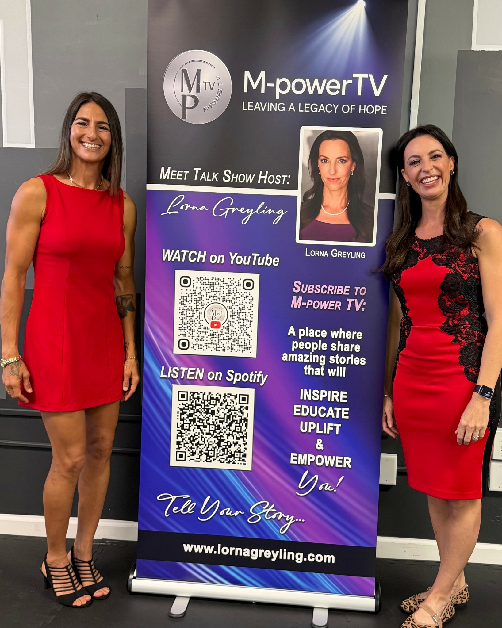 Two women in red dresses stand beside a vertical banner with text and QR codes, smiling indoors.
