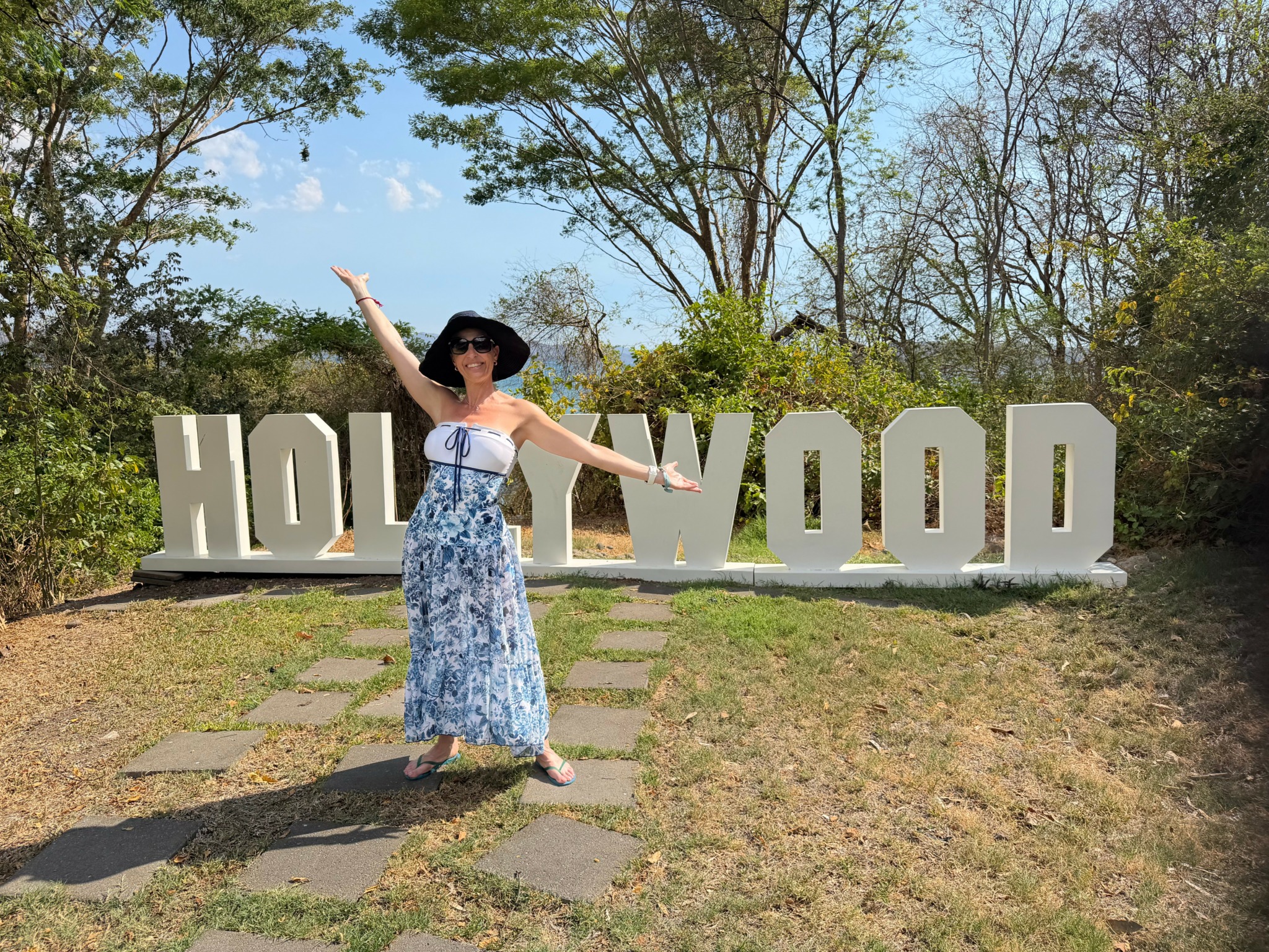 Person in a long dress and hat standing in front of Hollywood sign, arms raised, outdoors with trees and blue sky.