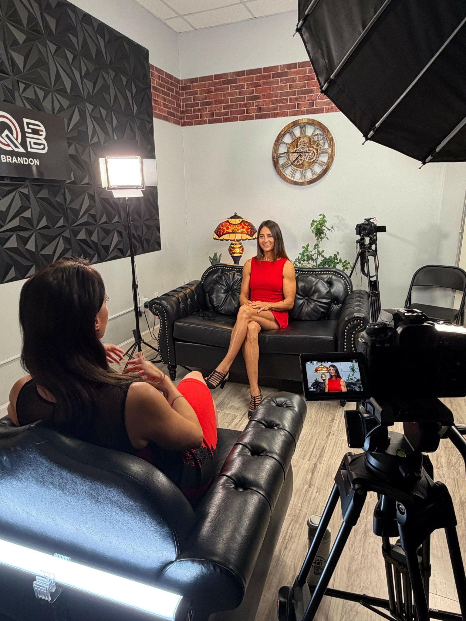 Woman in red dress and mask stands in a photography studio with equipment and a black leather couch.