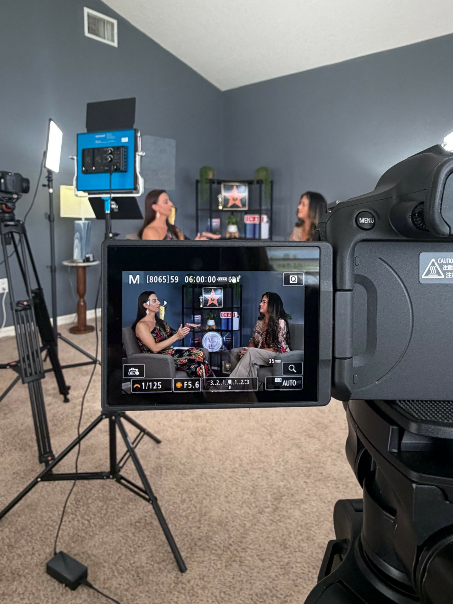 Camera viewfinder showing a person taking a photo in a room with gray walls and a tripod.