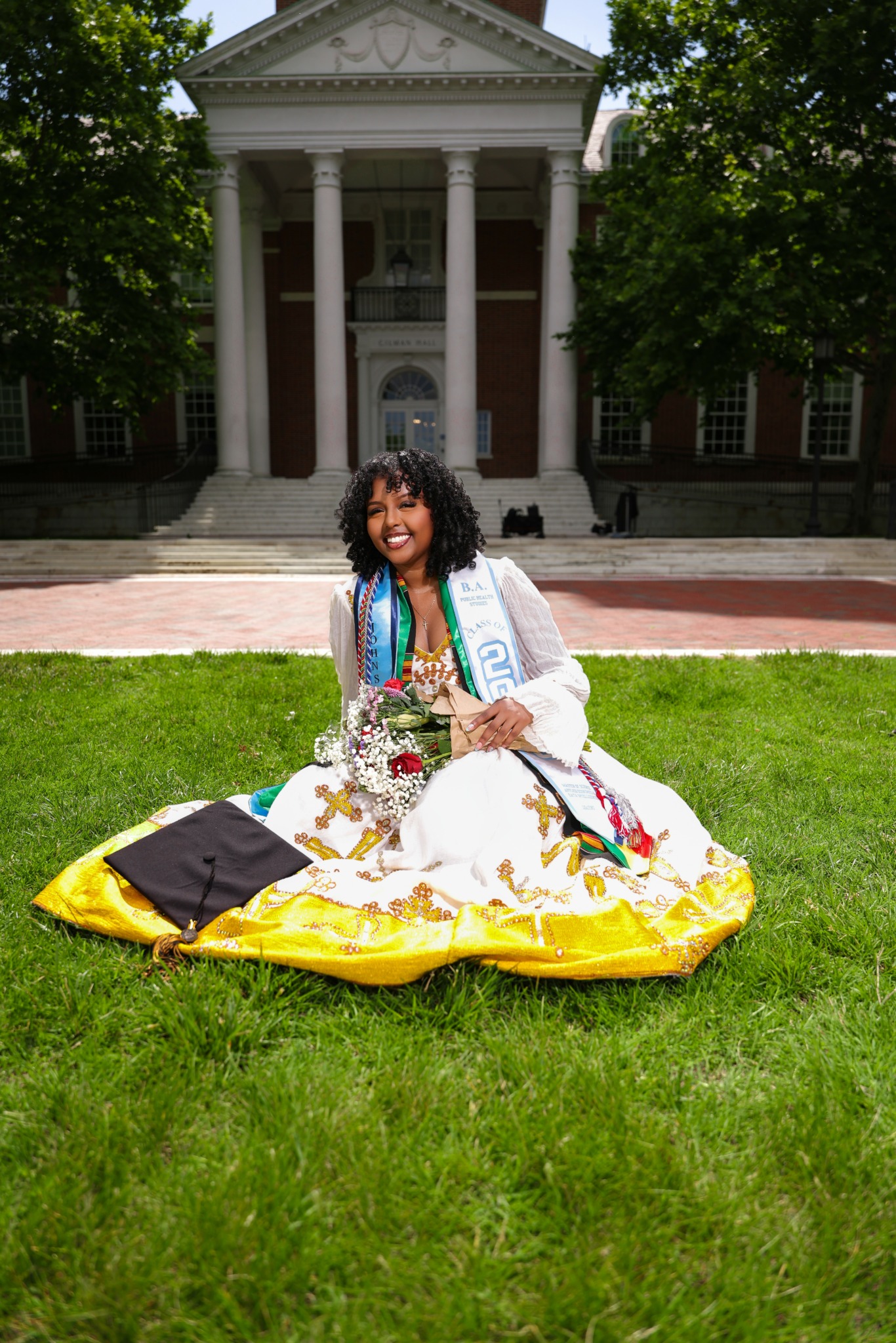 Young woman in graduation gown sitting on grass with diploma and flowers in front of a historic building with columns.