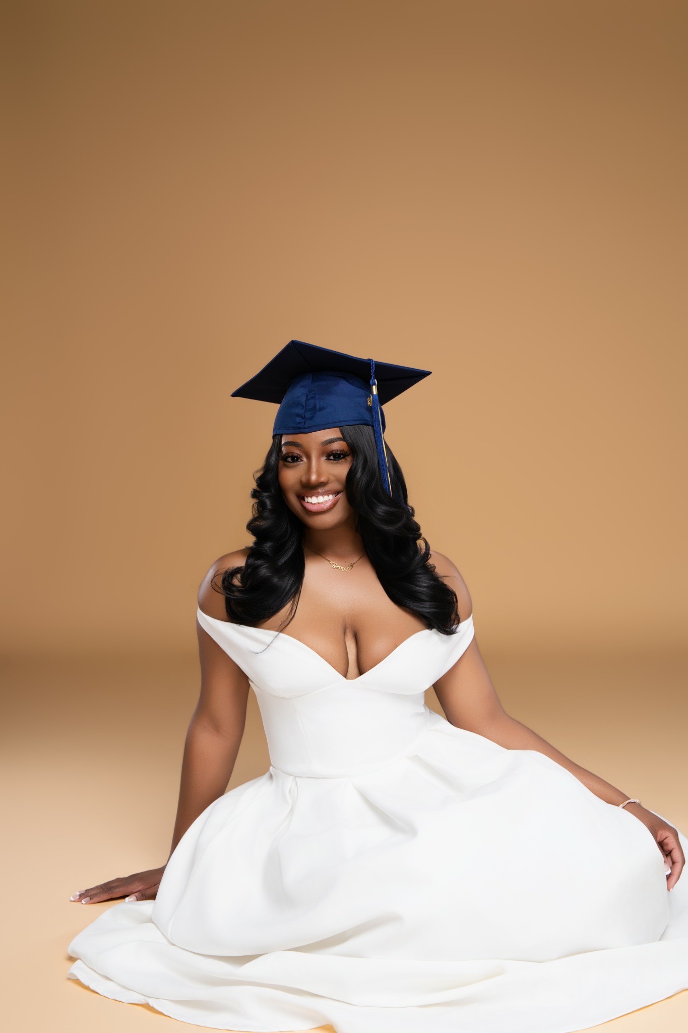 Woman in a white dress and graduation cap sitting on the floor smiling against a plain background.
