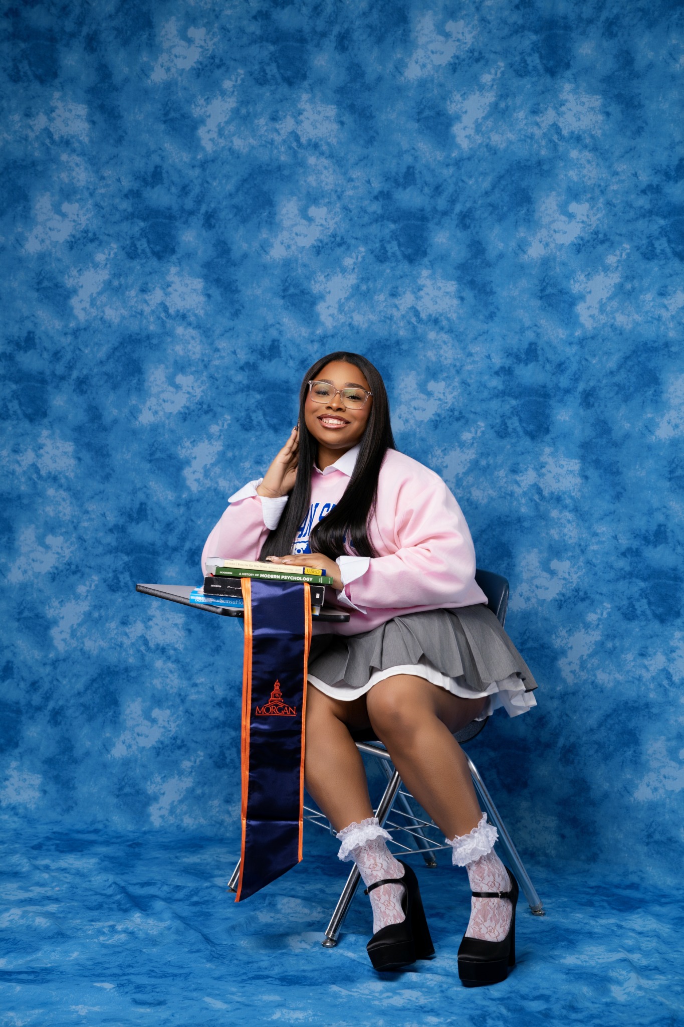 Young woman sitting on a chair with a blue textured background, smiling, wearing a pink jacket, gray skirt, and high heels.