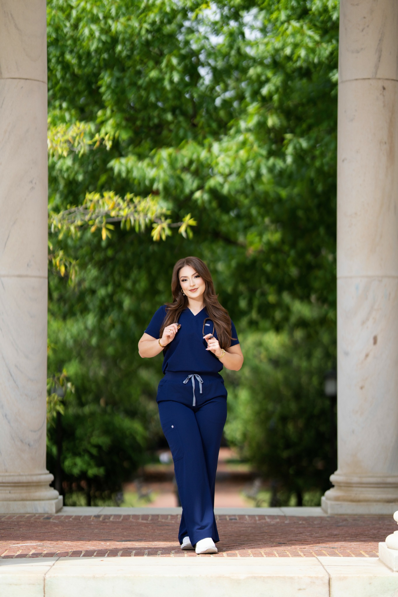 Young woman in navy outfit standing between two stone columns with green trees behind her.