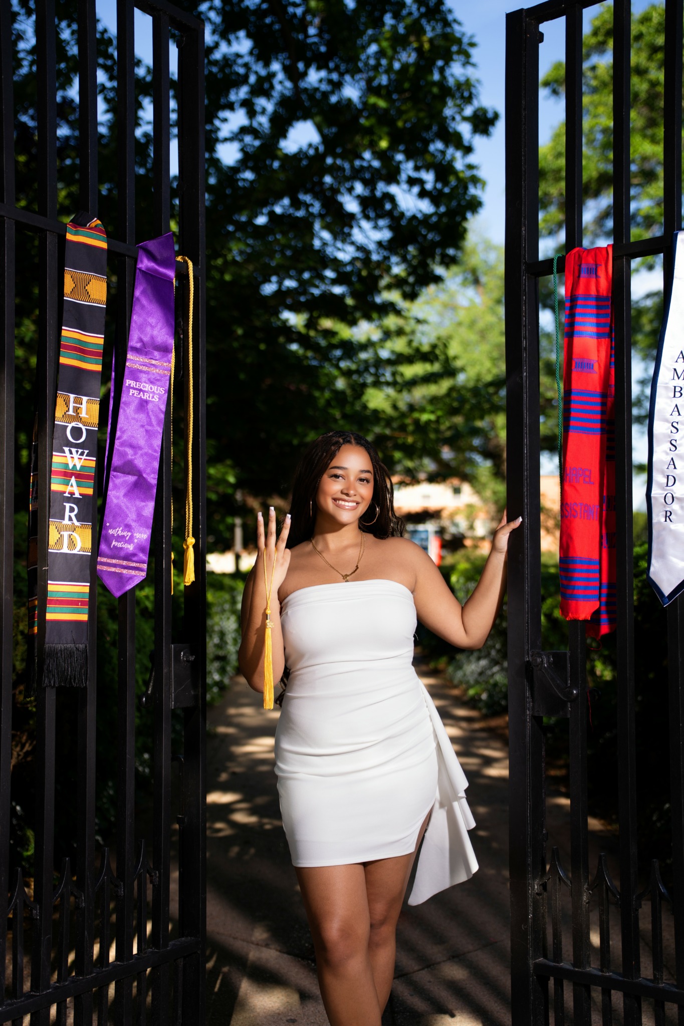 Woman in white dress standing between open gates with colorful sashes, smiling outdoors with trees and sunlight.