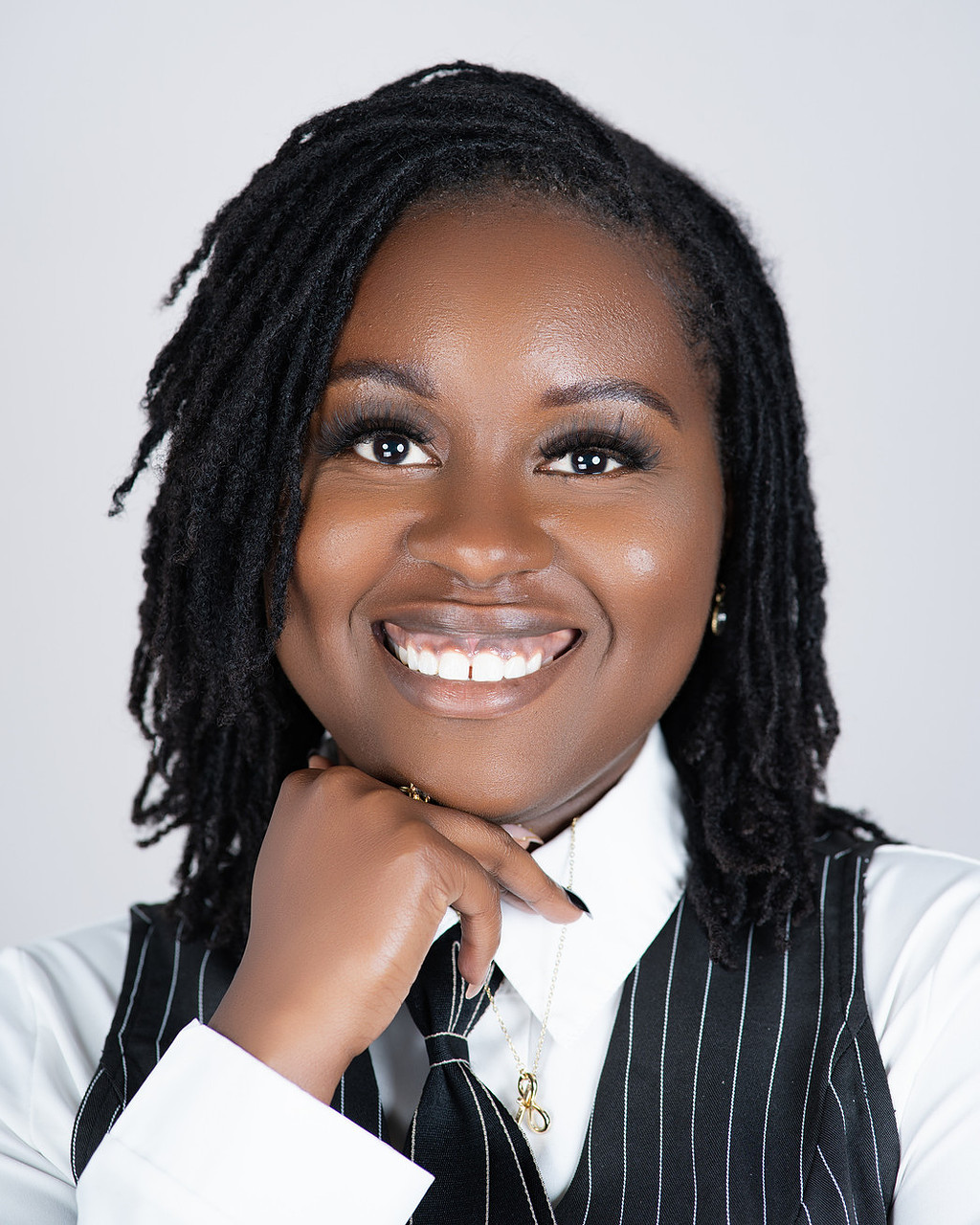 Smiling woman with dreadlocks, wearing a white shirt and black pinstripe vest, resting her chin on her hand.