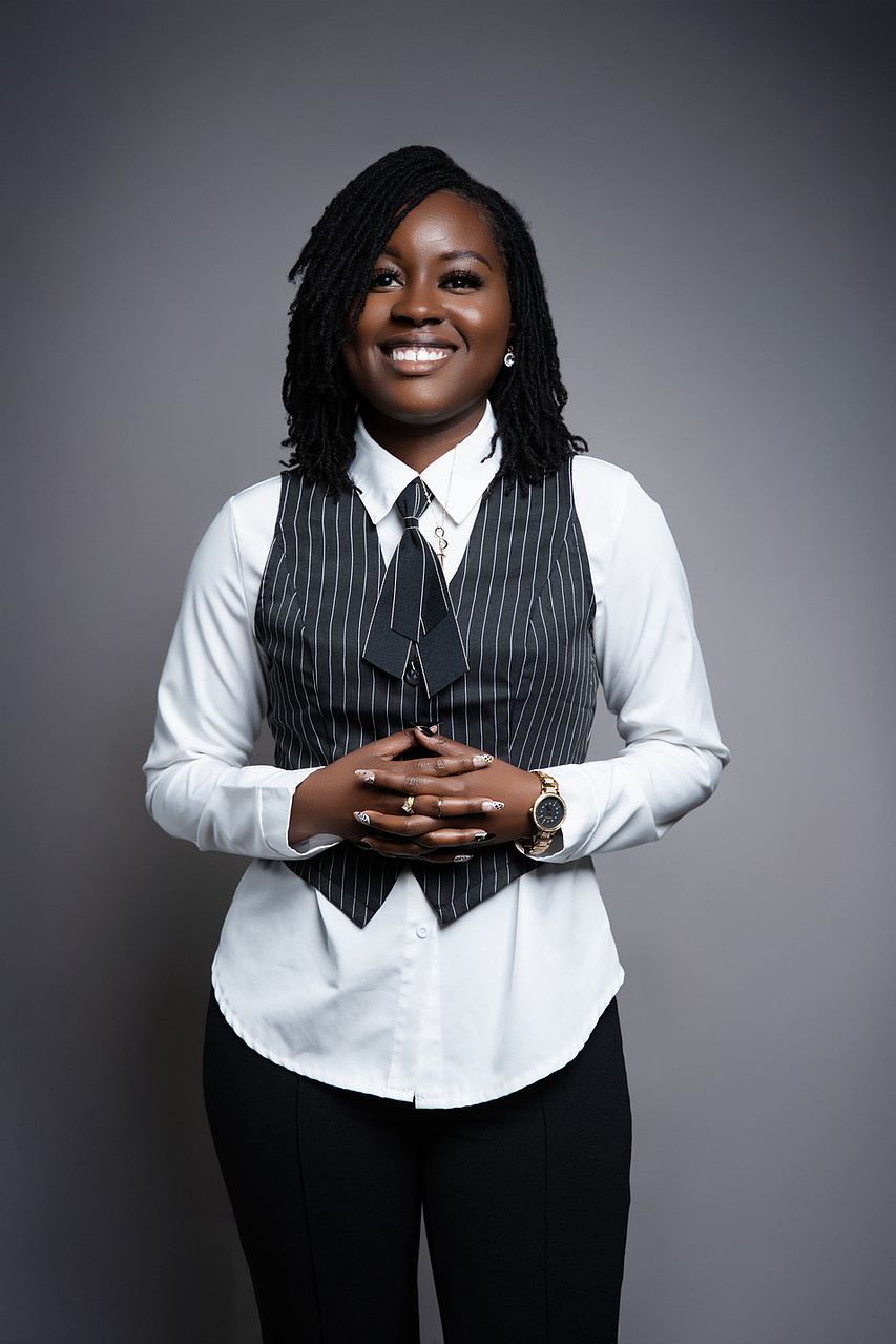 Woman with shoulder-length curly hair, smiling, wearing a white shirt, dark vest, and black pants, standing against a gray background.