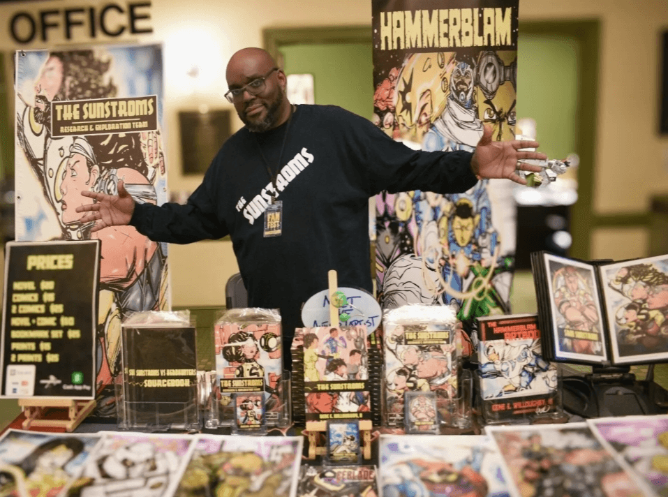 Person standing behind a table with comic books and artwork, smiling with arms outstretched in a store or convention setting.