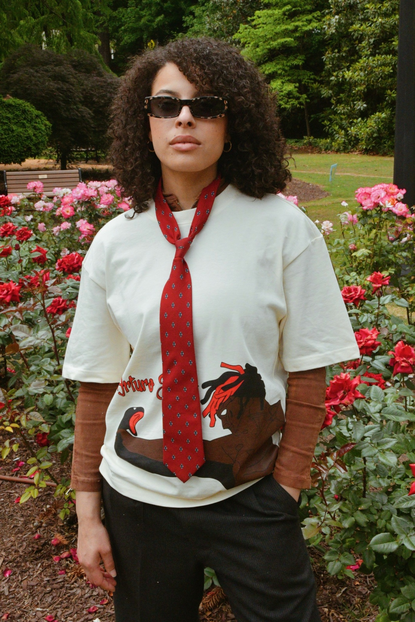 Woman with curly hair wearing sunglasses, a white shirt with horse print, red tie, and brown sleeves, standing in a garden with pink flowers.