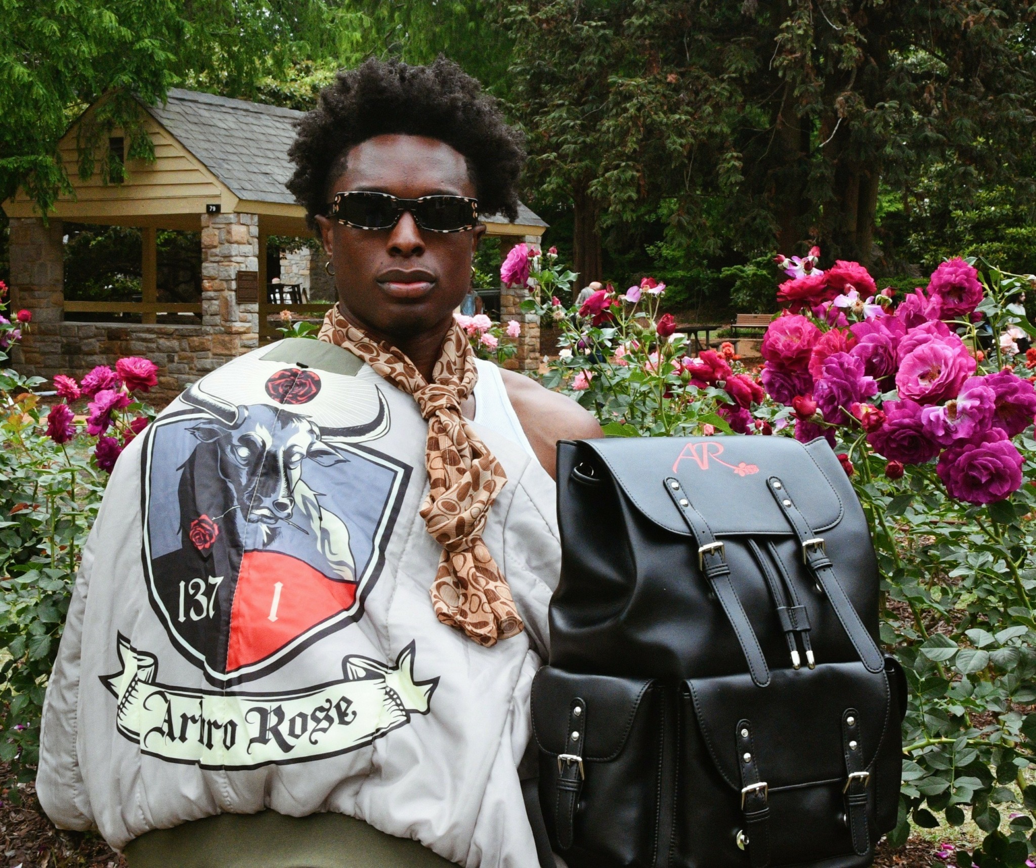 Person with sunglasses and scarf standing in garden with pink flowers, holding a black backpack, wearing a jacket with patches.
