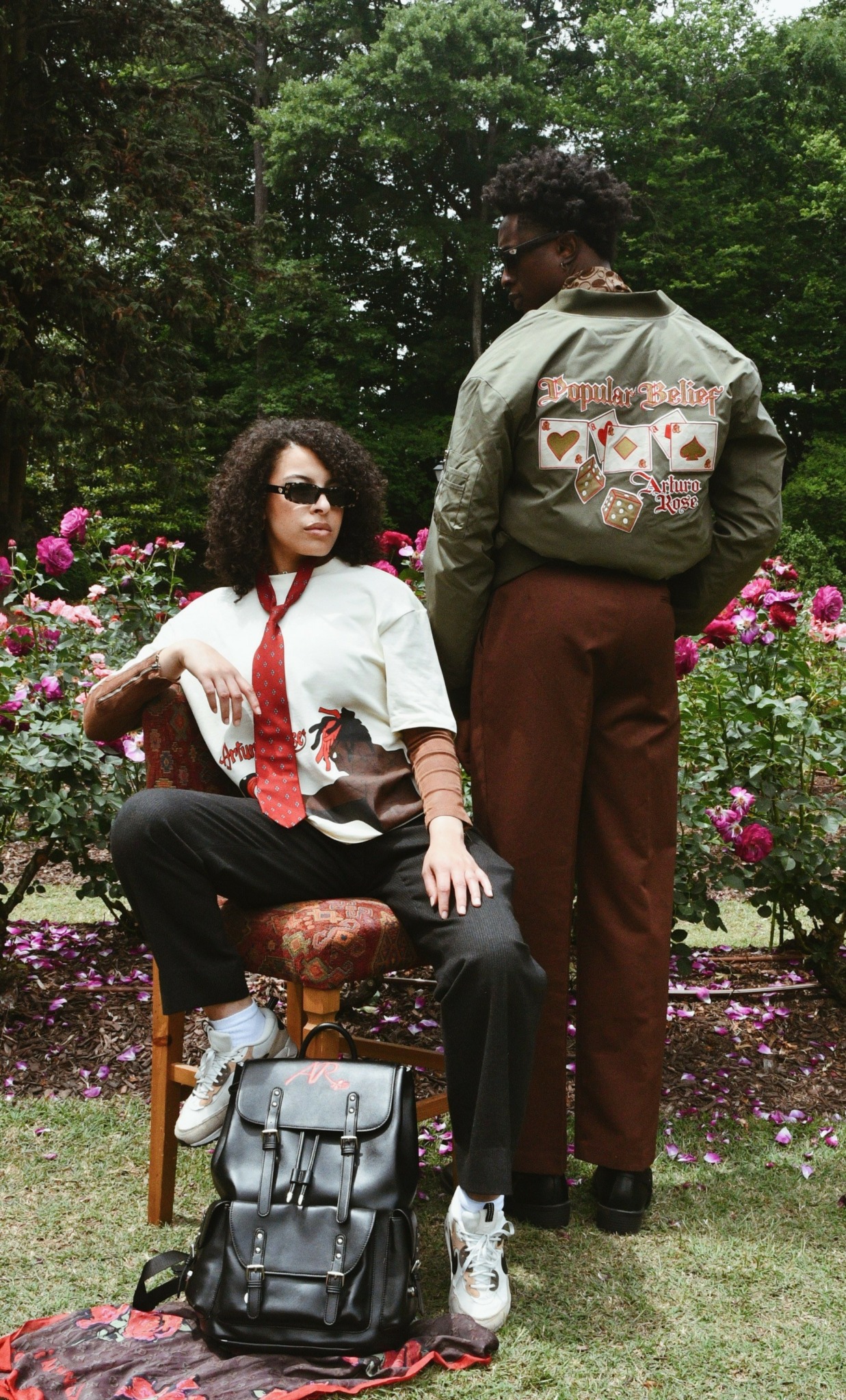 Two women in a garden with pink flowers and green trees, one sitting on a chair and the other standing nearby.