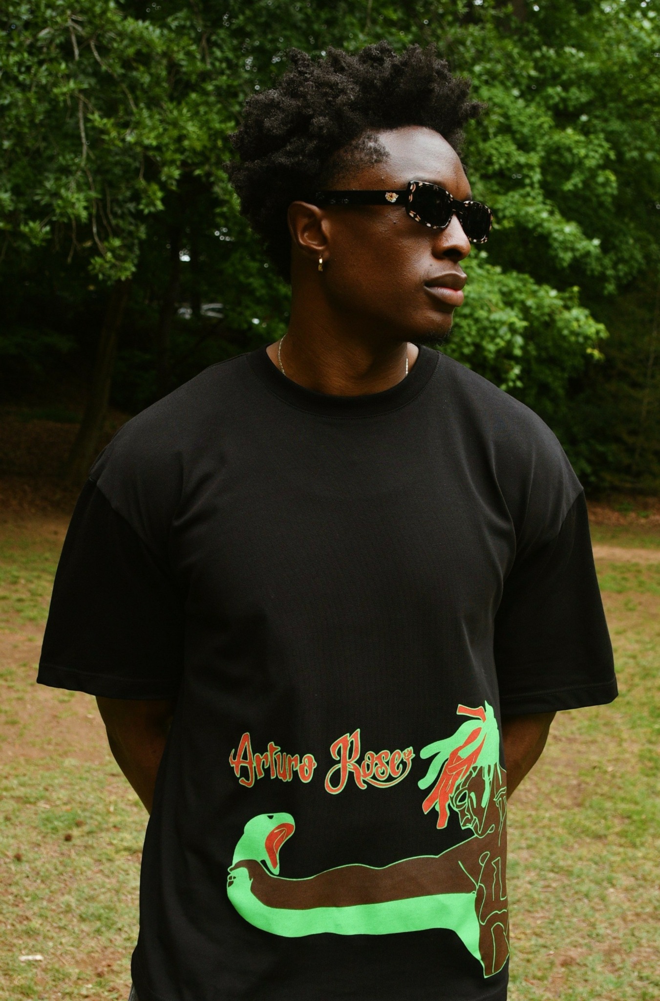 Woman with short curly hair wearing sunglasses and a black T-shirt outdoors, trees in background.