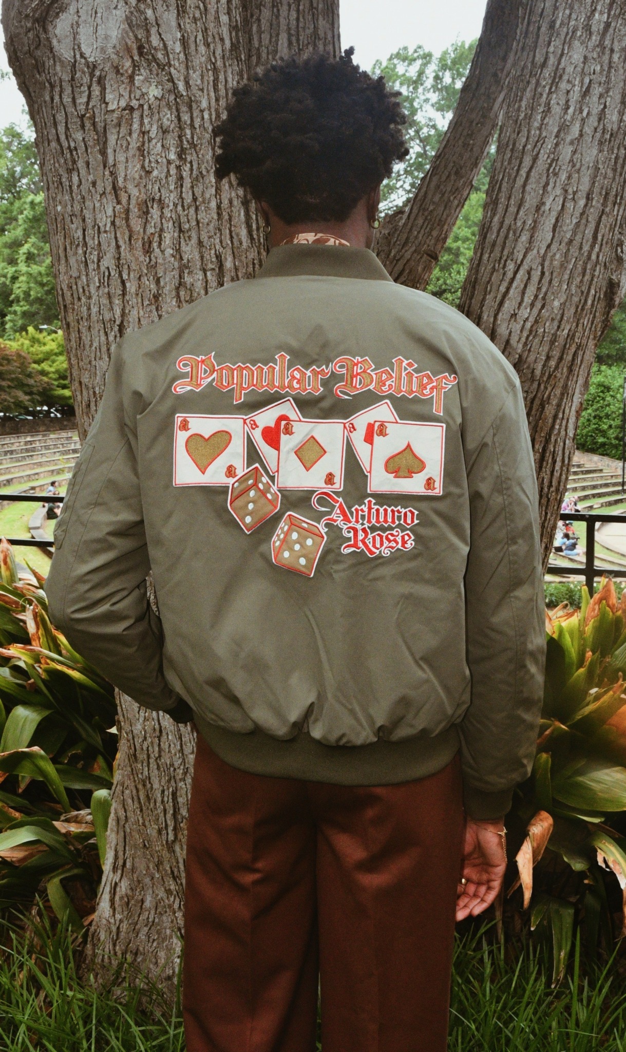 Person with curly hair wearing a green jacket with playing card graphics, standing outdoors near trees and plants.