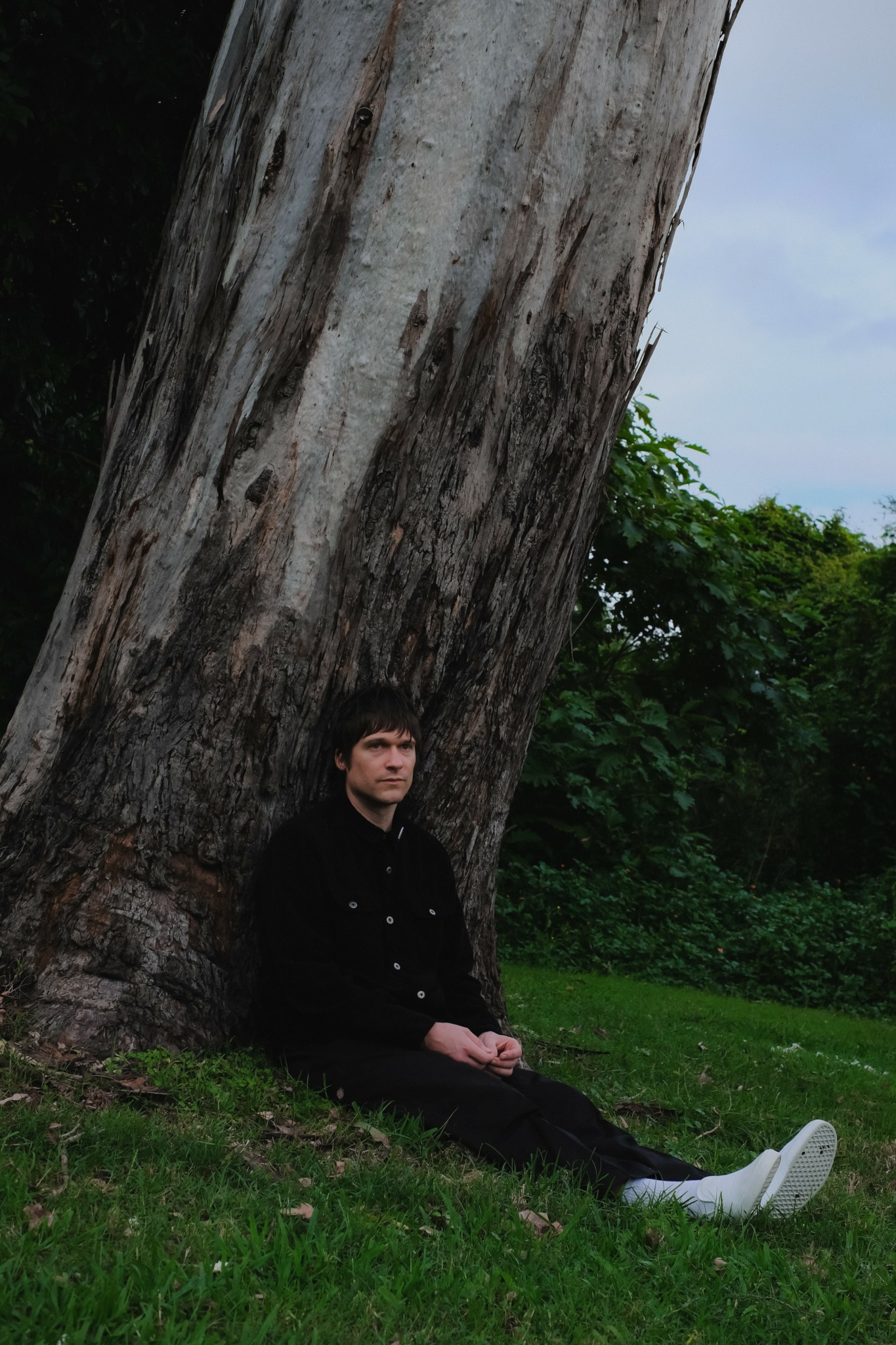 Person sitting on grass leaning against a large tree trunk outdoors, with greenery and sky in background.