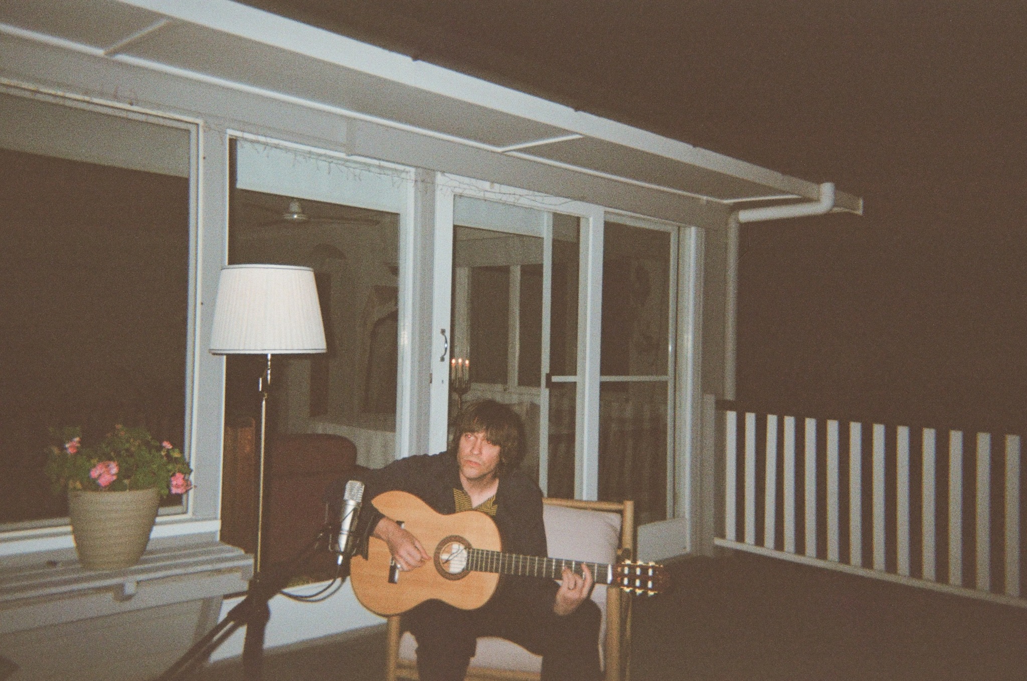 Person sitting on a chair playing an acoustic guitar on a balcony at night, with a lamp and potted plant nearby.