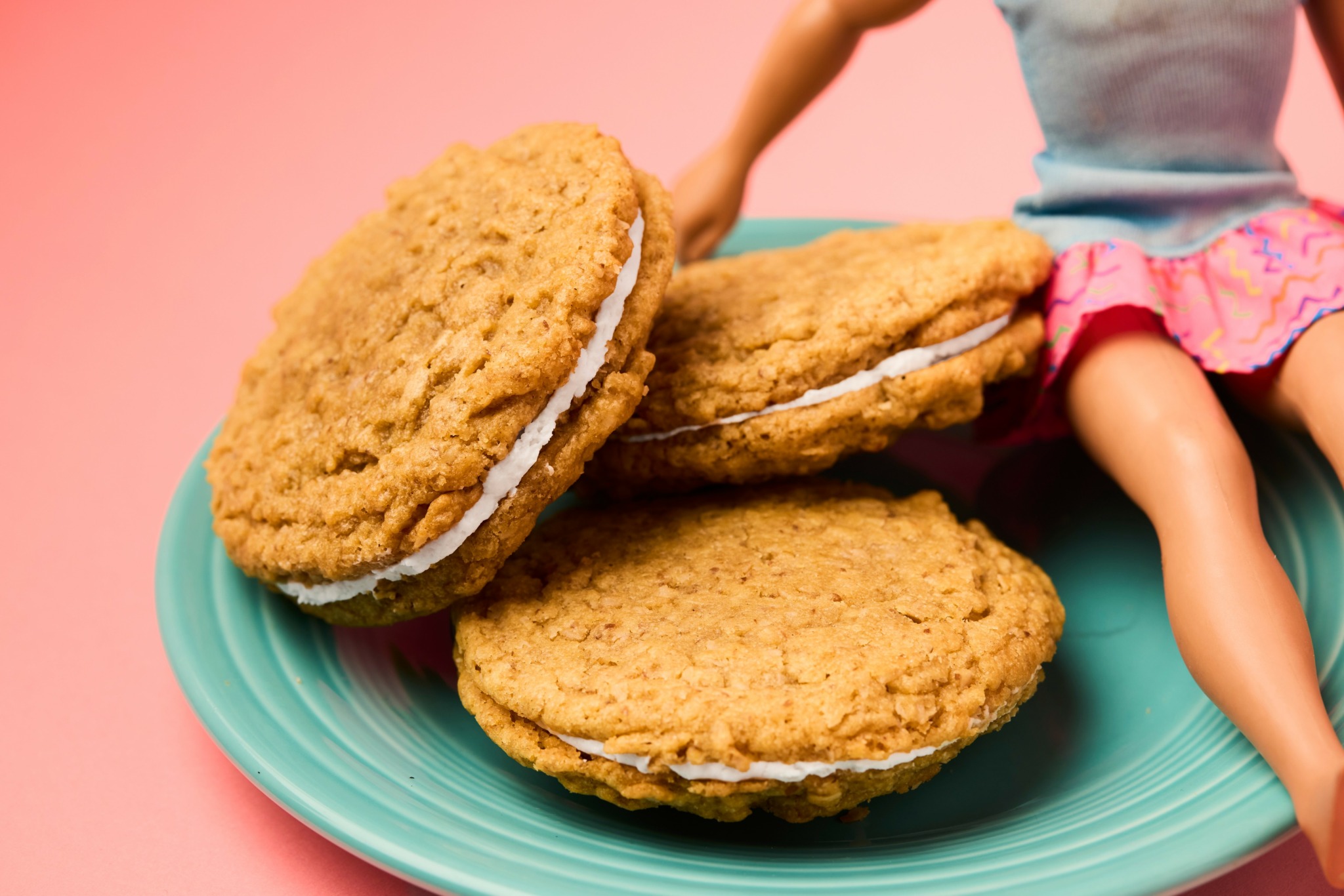 Three large oatmeal cookies on a teal plate with a doll sitting beside them, against a pink background.