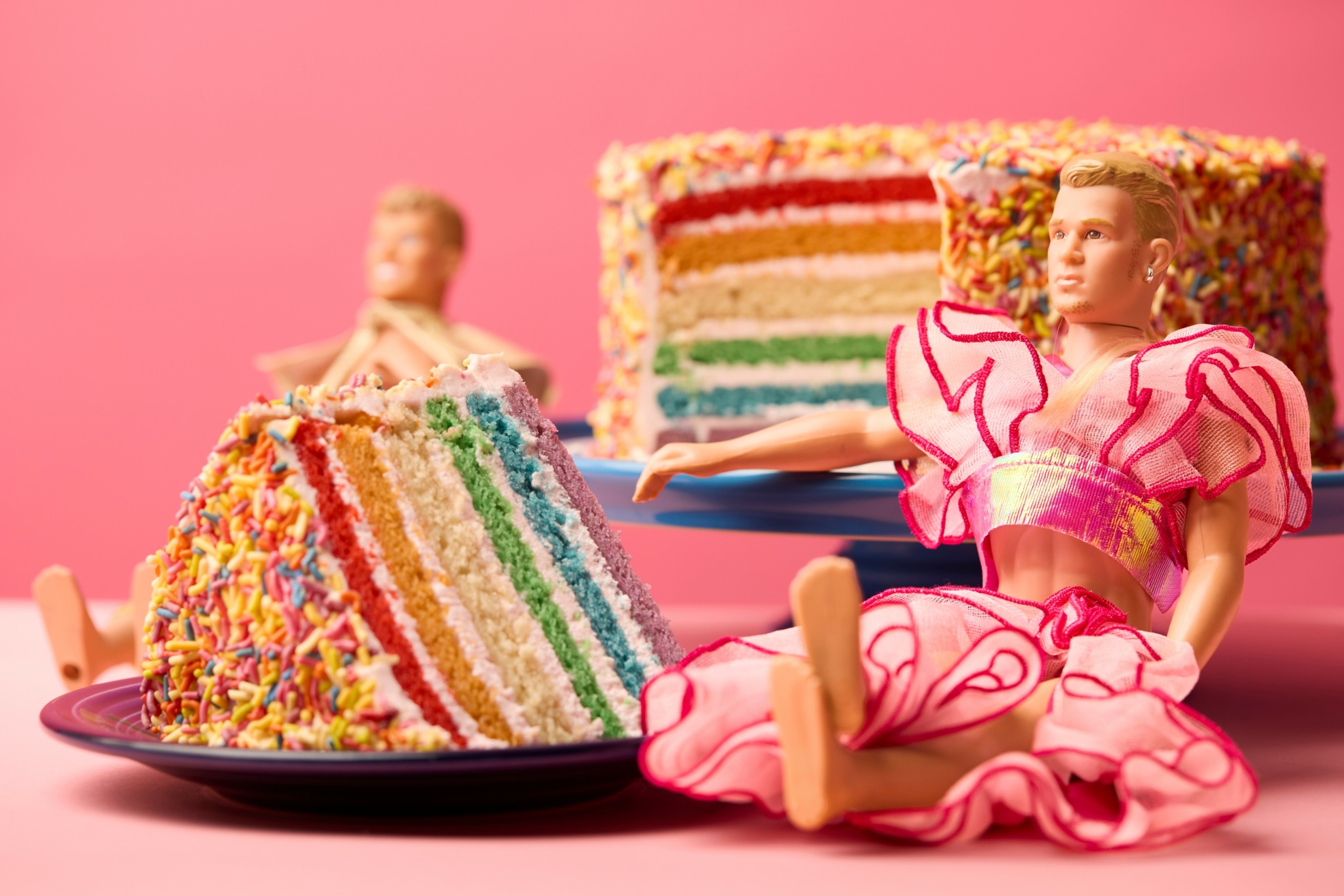 Two dolls sit beside a rainbow-colored cake with a slice removed, set against a pink background.