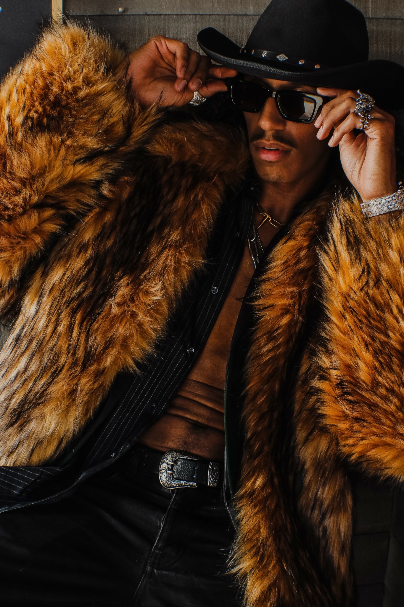 Person wearing a large fur coat, black hat, sunglasses, and jewelry, adjusting their hat with one hand.