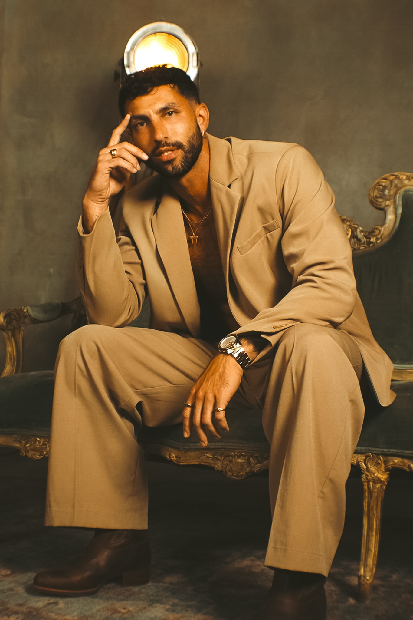 Man in beige suit sitting on a vintage sofa, resting his chin on his hand, with a dark background and a decorative lamp behind him.