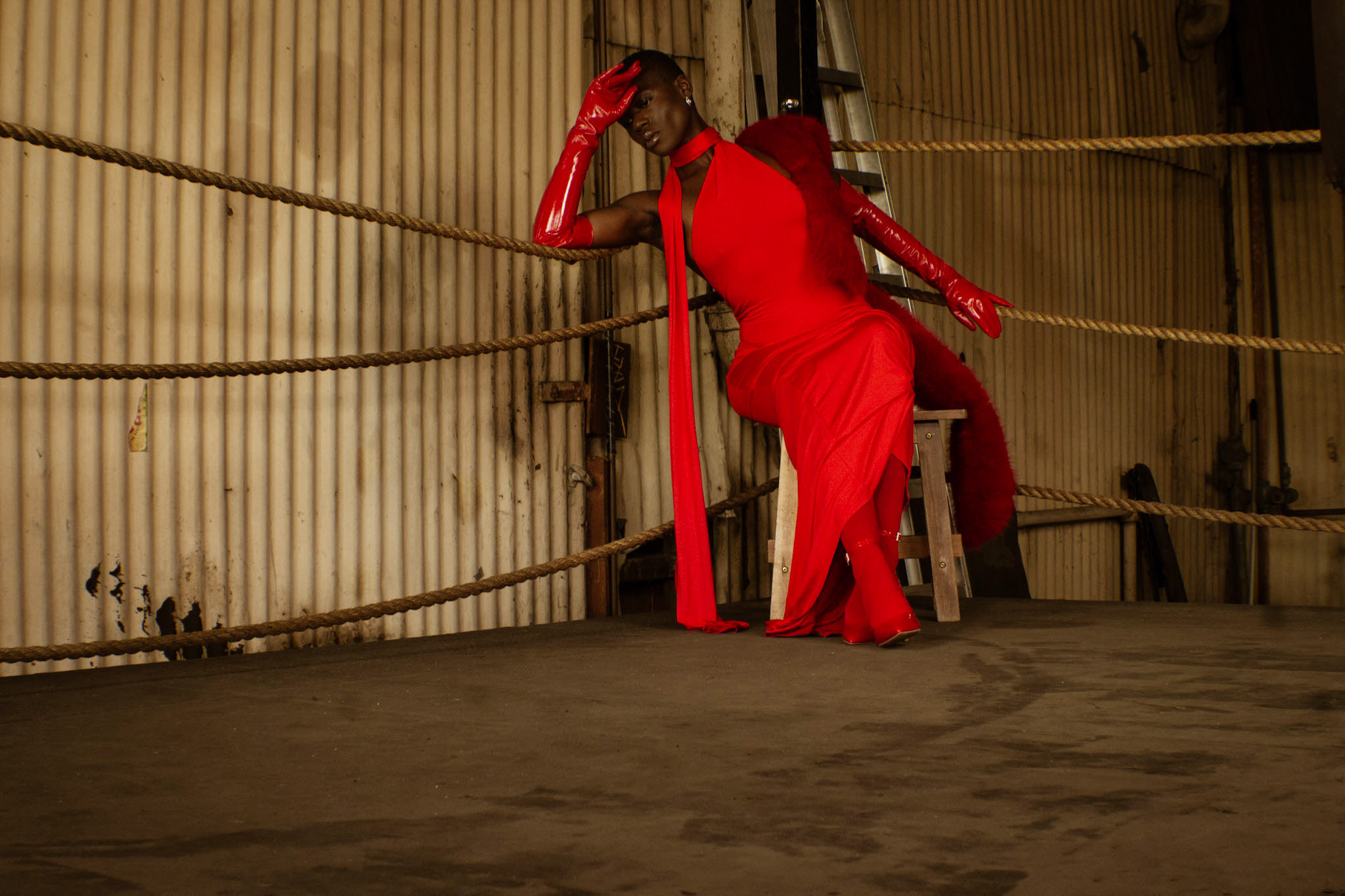 Person in a red dress and gloves leaning against boxing ring ropes in an industrial setting.