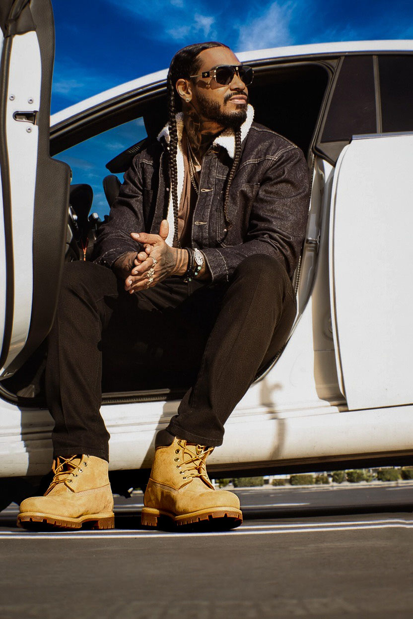Man with sunglasses and dreadlocks sitting in a white vehicle's open door, wearing a black jacket, black pants, and tan boots.
