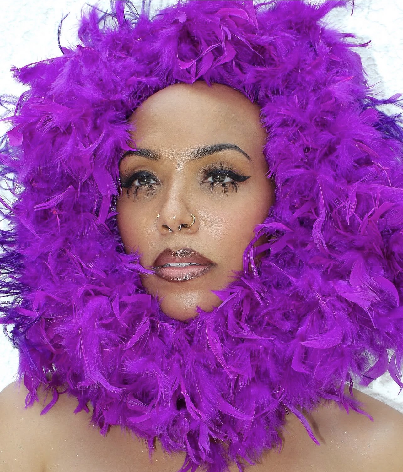 Person with purple feathered headpiece surrounding face, makeup, and nose piercing, looking at camera.