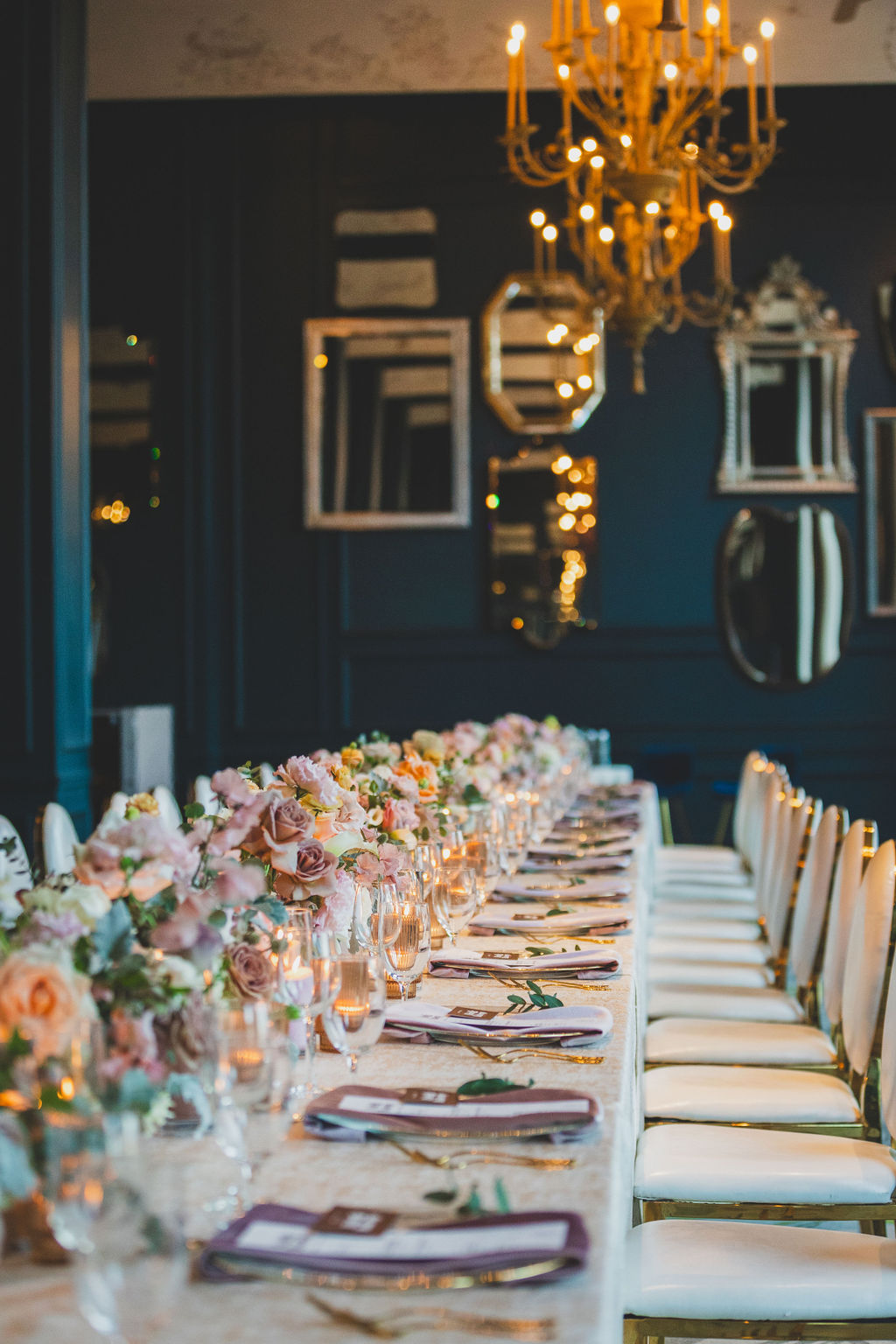 Long dining table set with floral arrangements, plates, and cutlery in an elegant room with chandeliers and framed mirrors.