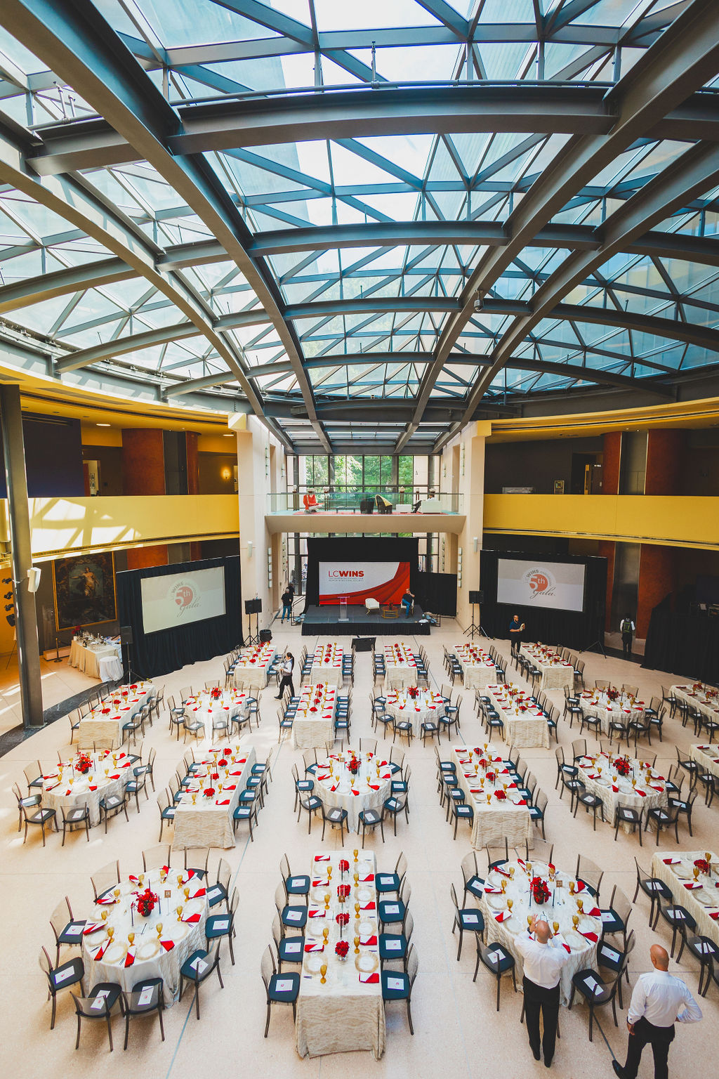 Event space with round tables, chairs, and a stage, under a glass ceiling with metal framework, set for a formal gathering.