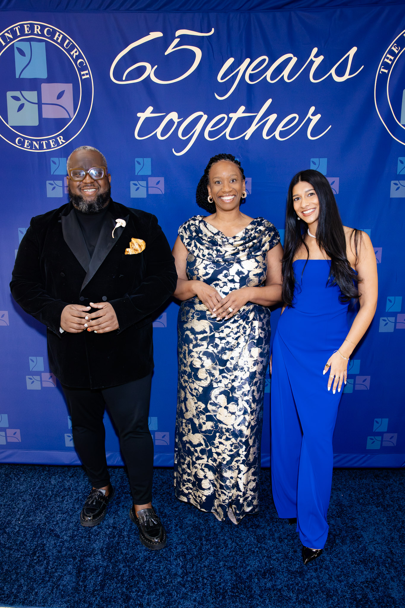 Three people standing in front of a blue backdrop with text '65 years together' and logos, smiling at the camera.