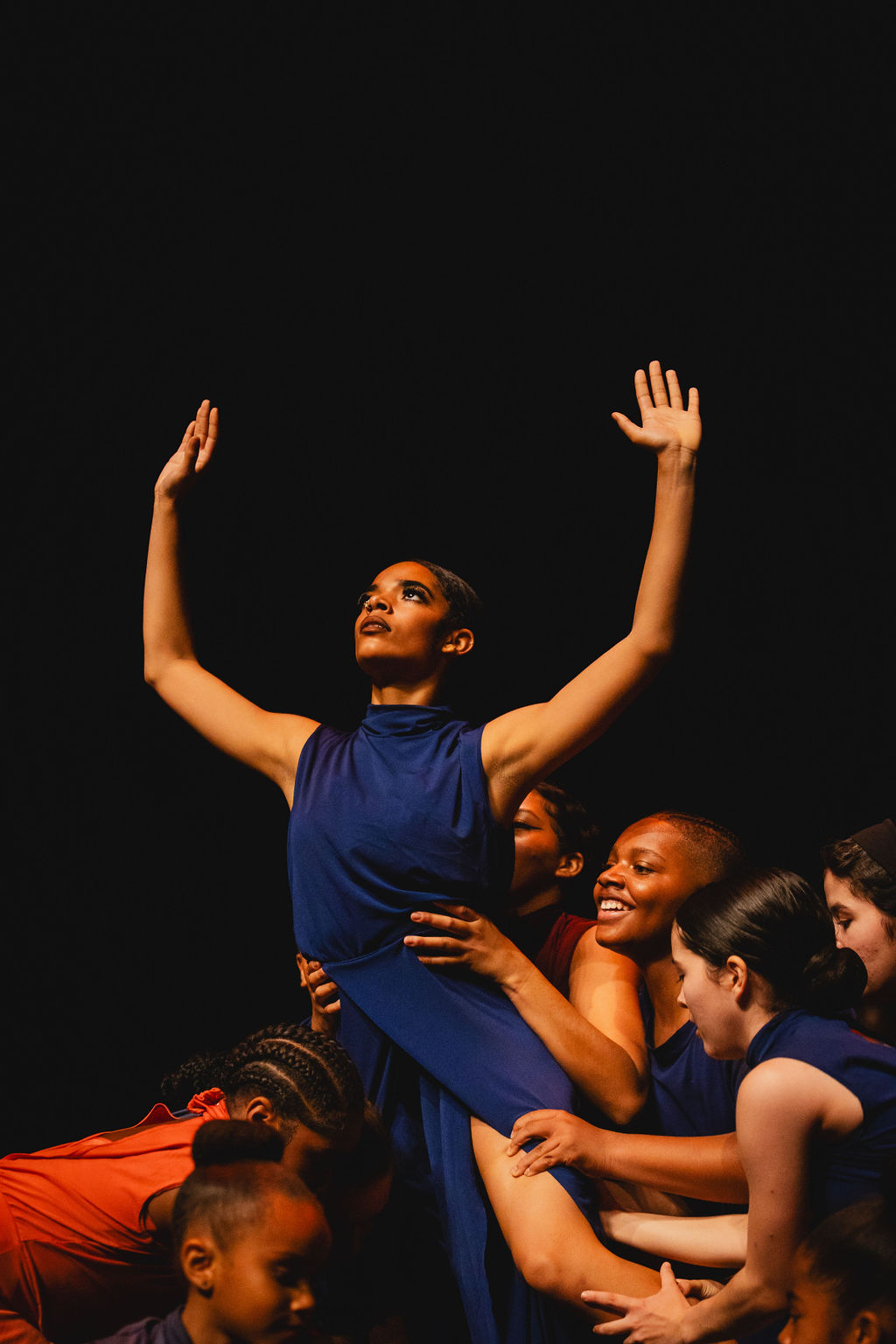 Group of women and children, with one woman in a blue dress raising her arms, against a black background.