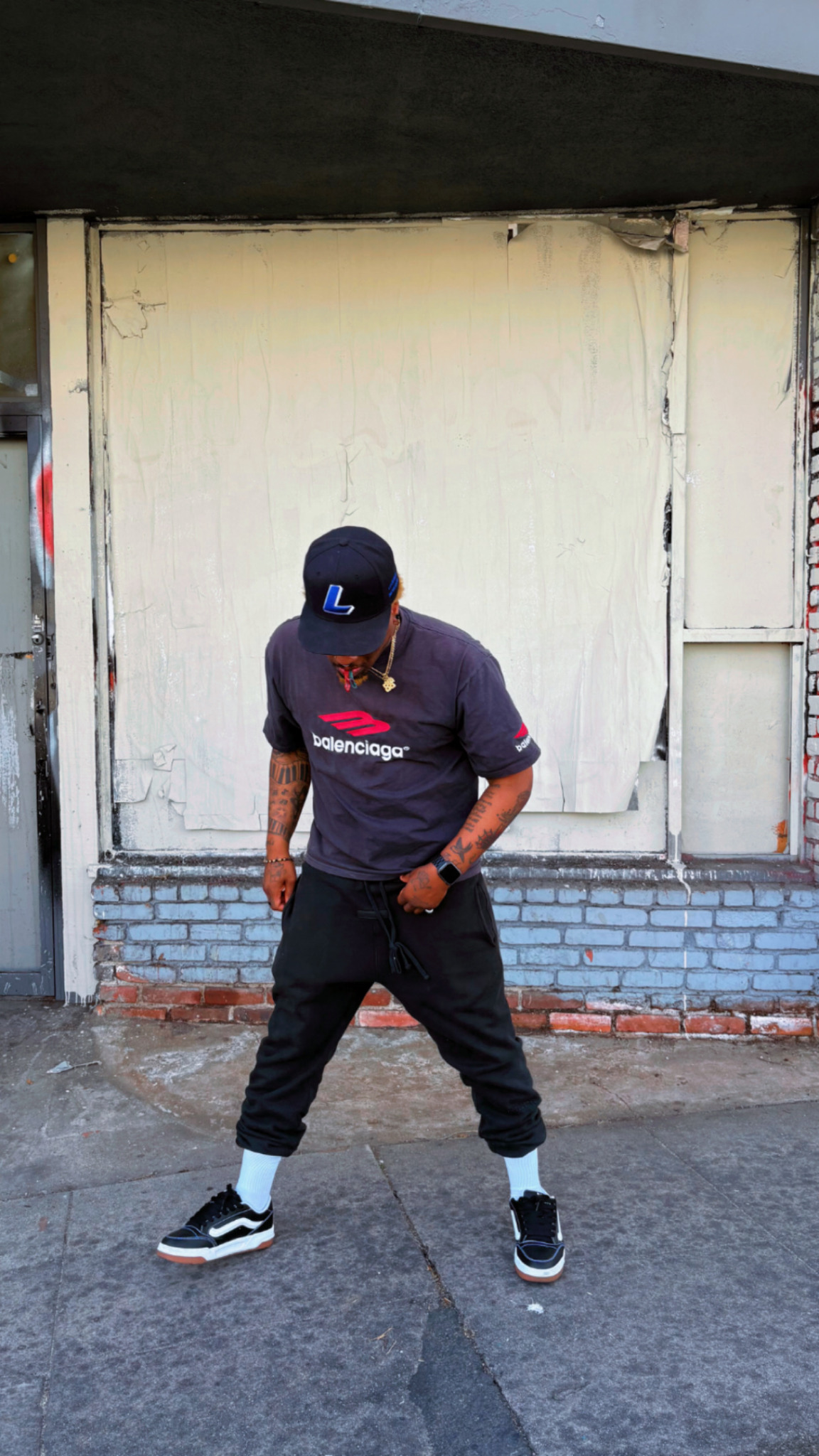Person standing outdoors in front of a wall, wearing a cap, t-shirt, and sneakers, with legs apart and hands on hips.