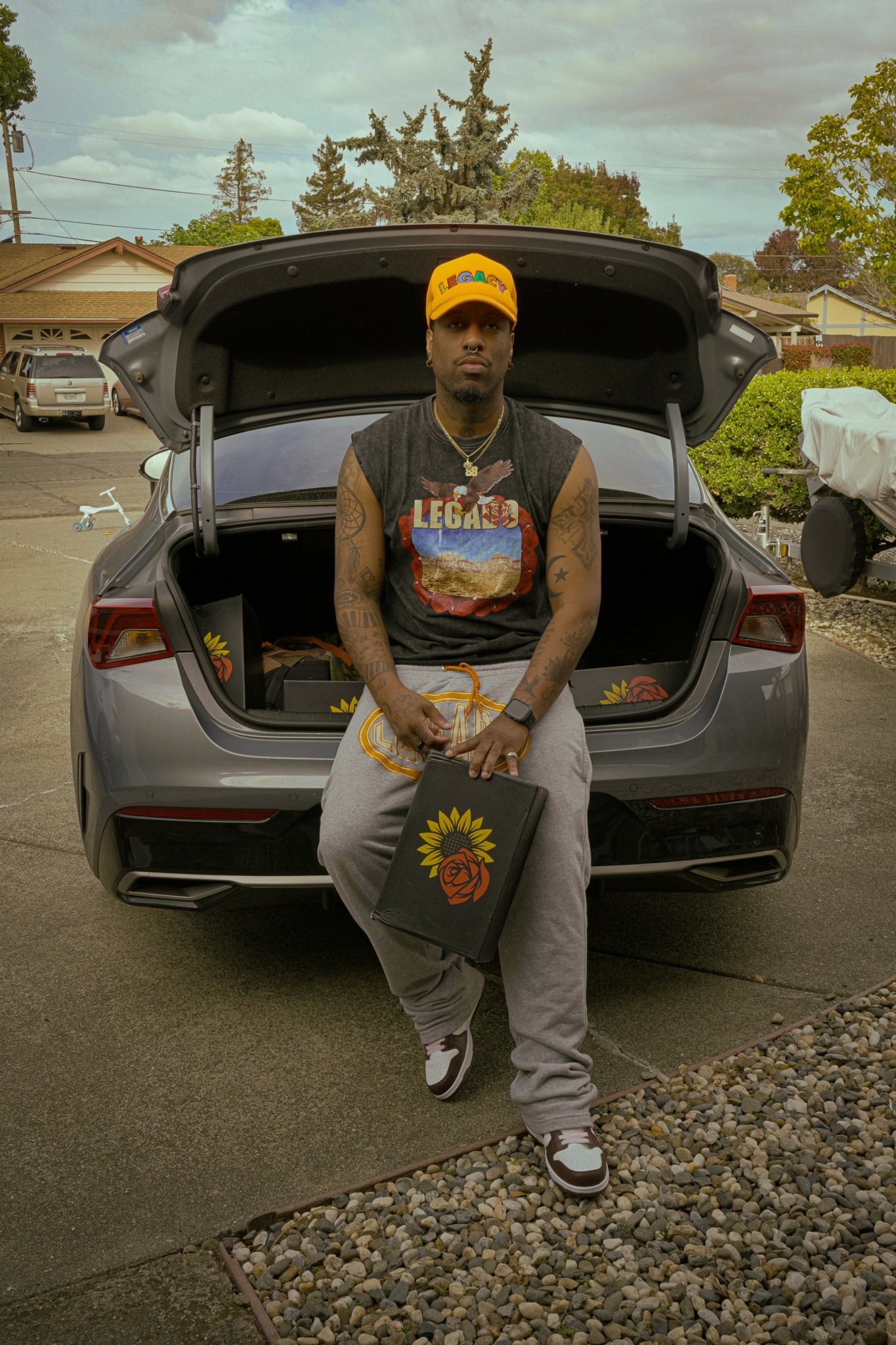 Young man with tattoos wearing a yellow cap and gray pants, sitting on a car's open trunk in a residential neighborhood.