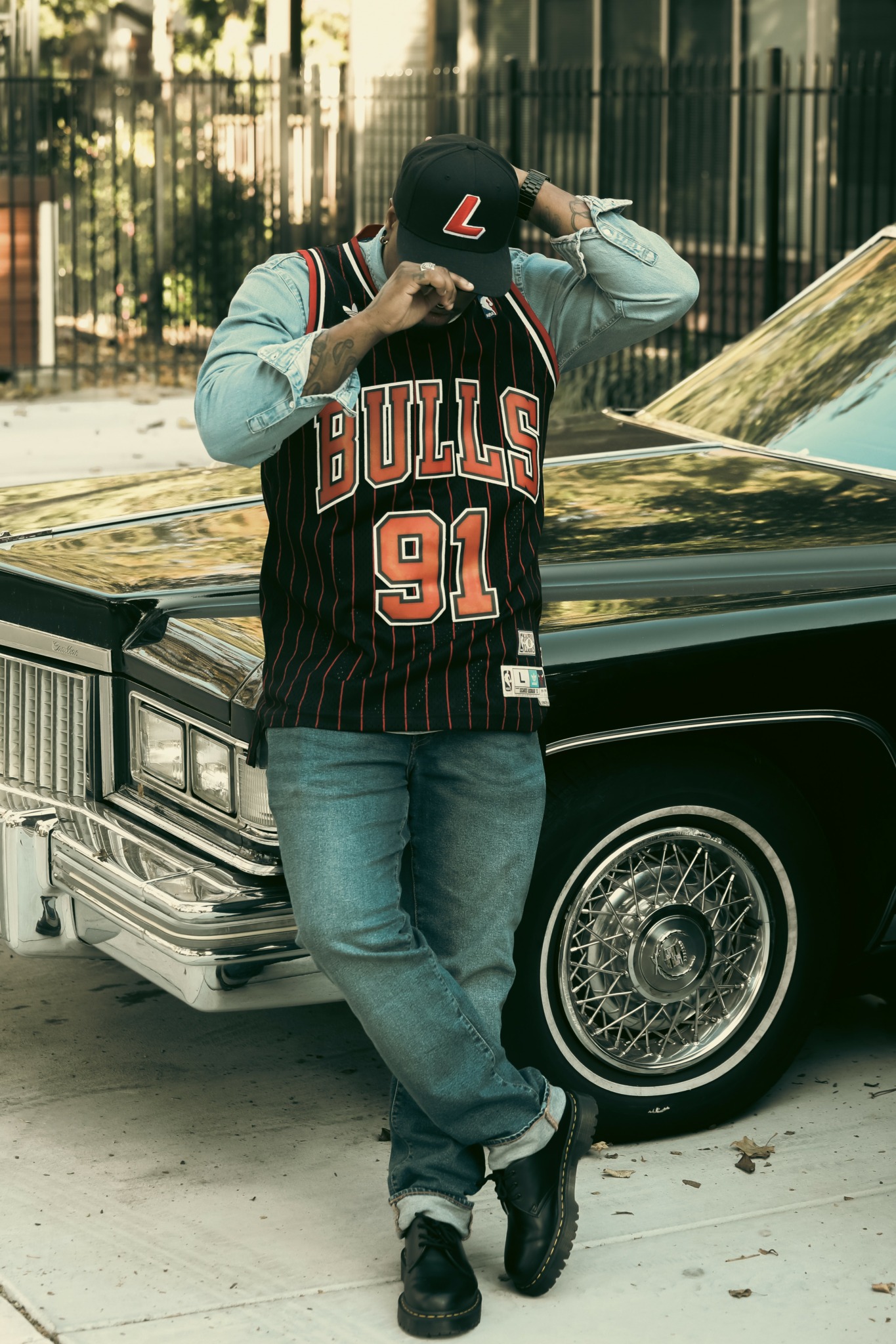 Person wearing a Chicago Bulls jersey and cap, standing next to a black vintage car, adjusting their cap with one hand.