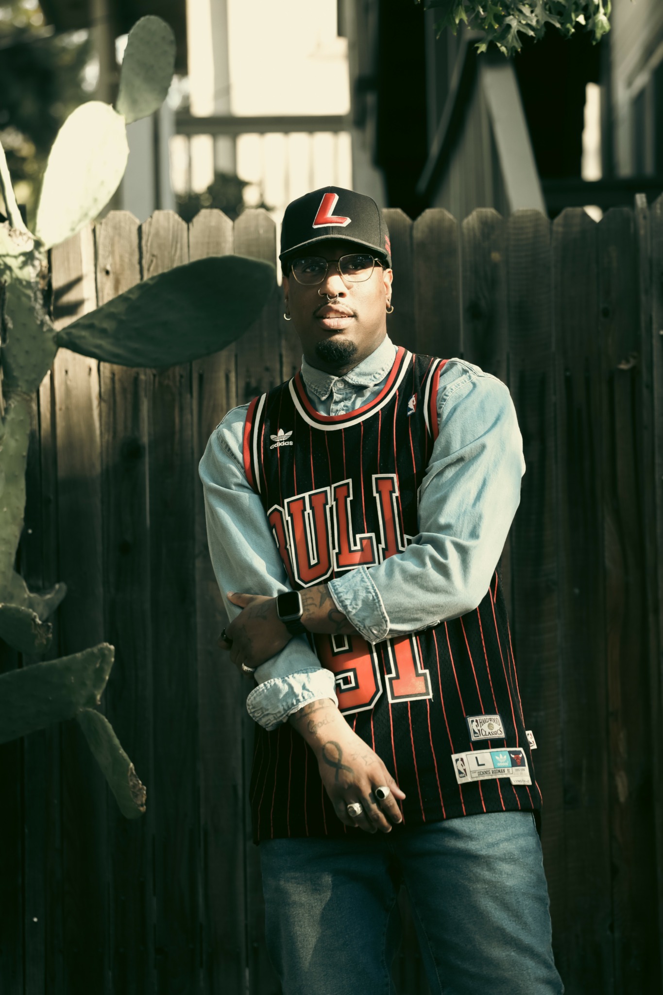 Young man wearing a Chicago Bulls jersey, cap, and sunglasses, standing outdoors near a wooden fence and cactus.