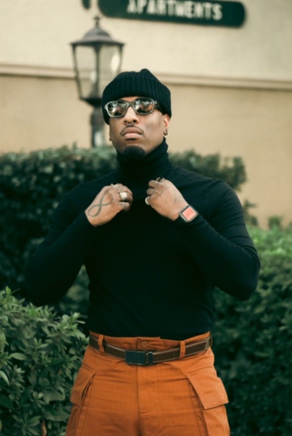 Person wearing black beanie, sunglasses, black turtleneck, and orange pants standing outdoors with greenery and a lamp post behind.