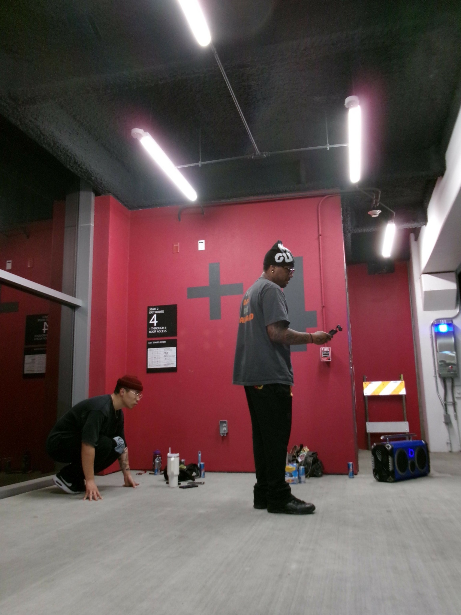 Two people in a room with a red wall, one standing and one kneeling, with various items on the floor.