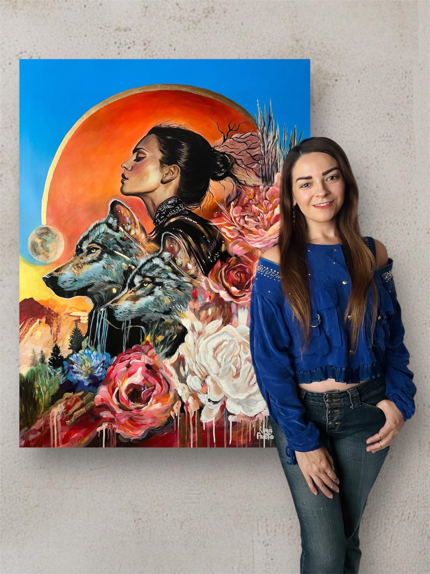 Young woman with long brown hair wearing a blue jacket and jeans, standing next to a colorful painting of a woman, wolf, and flowers.