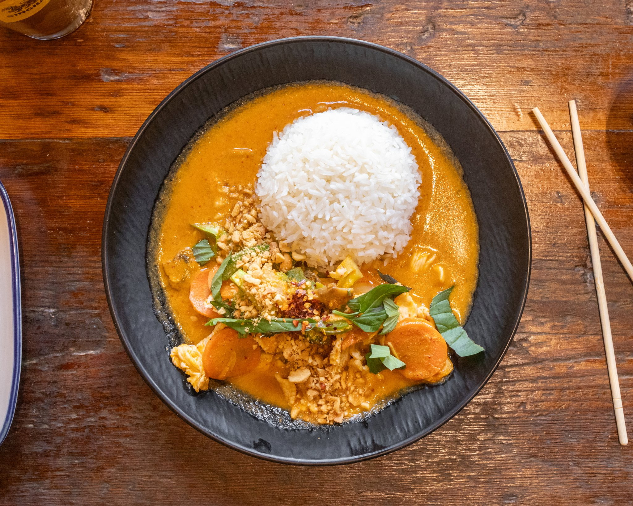 Bowl of rice with curry, garnished with basil leaves on a wooden table, chopsticks beside the bowl.