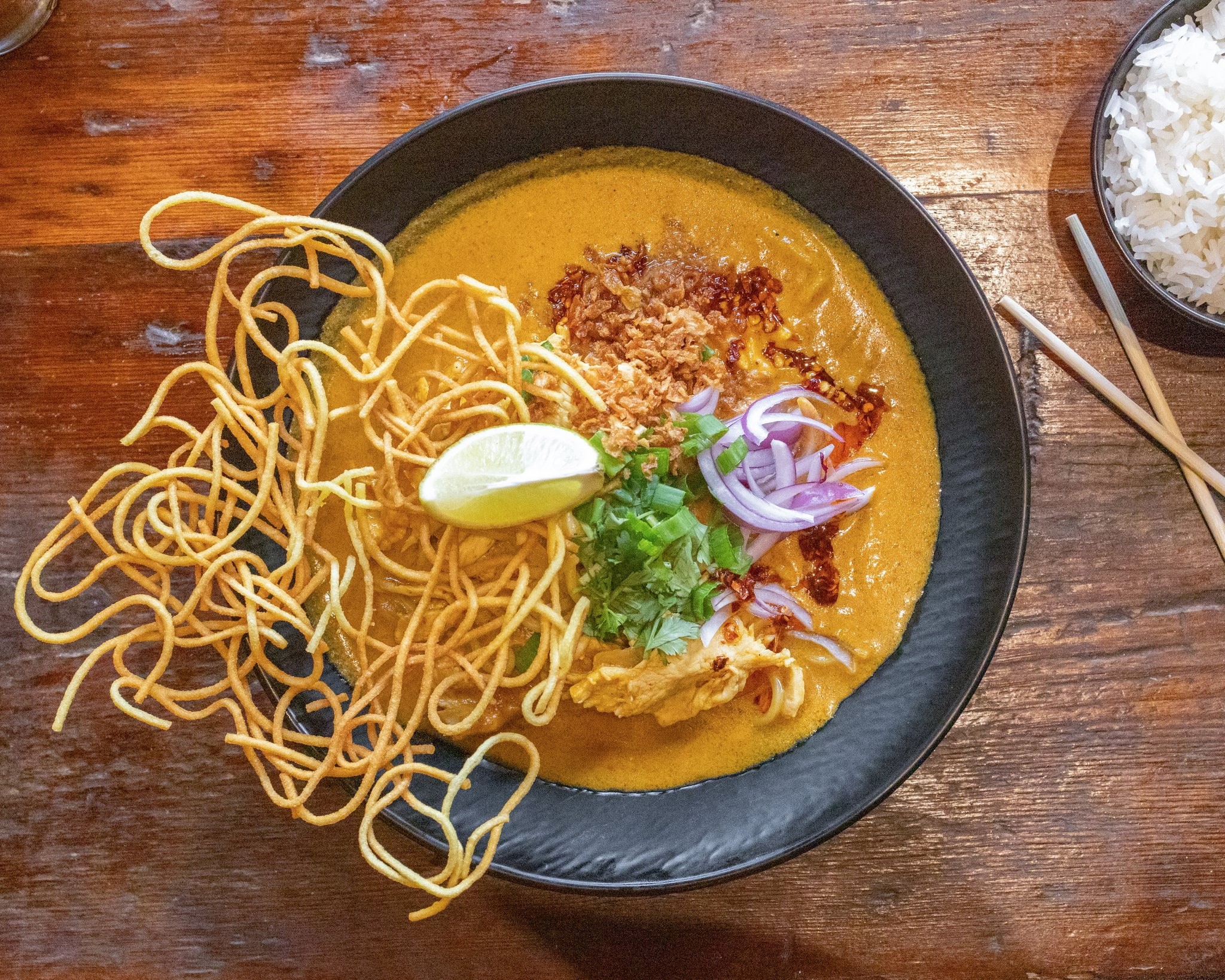 Bowl of yellow curry with noodles, lime wedge, red onion, and cilantro on a wooden table, with rice in a side bowl