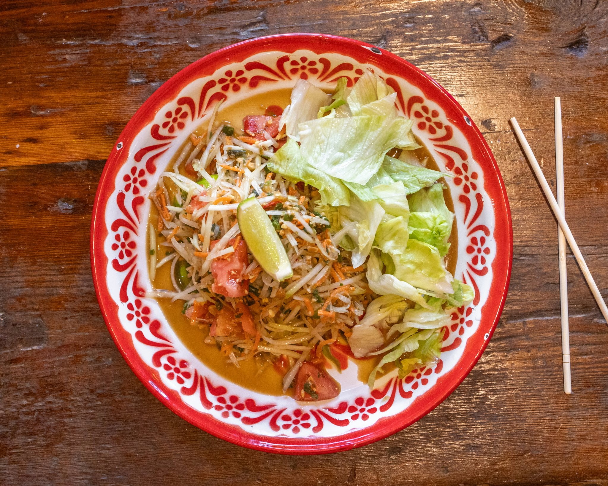 Bowl of noodle salad with lettuce, lime wedge, and chopsticks on wooden table.