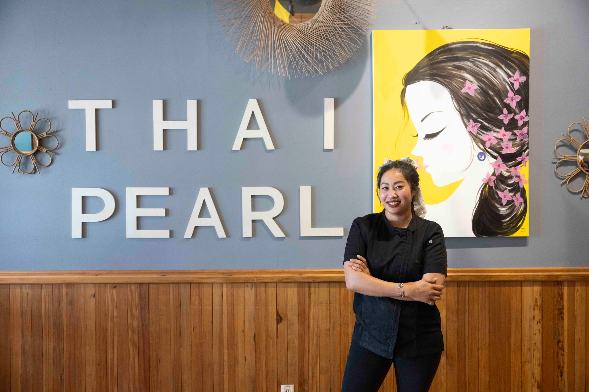 Woman standing with arms crossed in front of a wall with Thai Pearl sign and artwork, smiling.