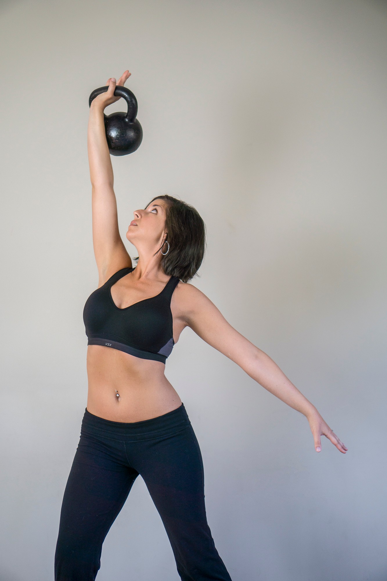 Woman in workout clothes lifting a kettlebell overhead with one arm, standing against a plain background.