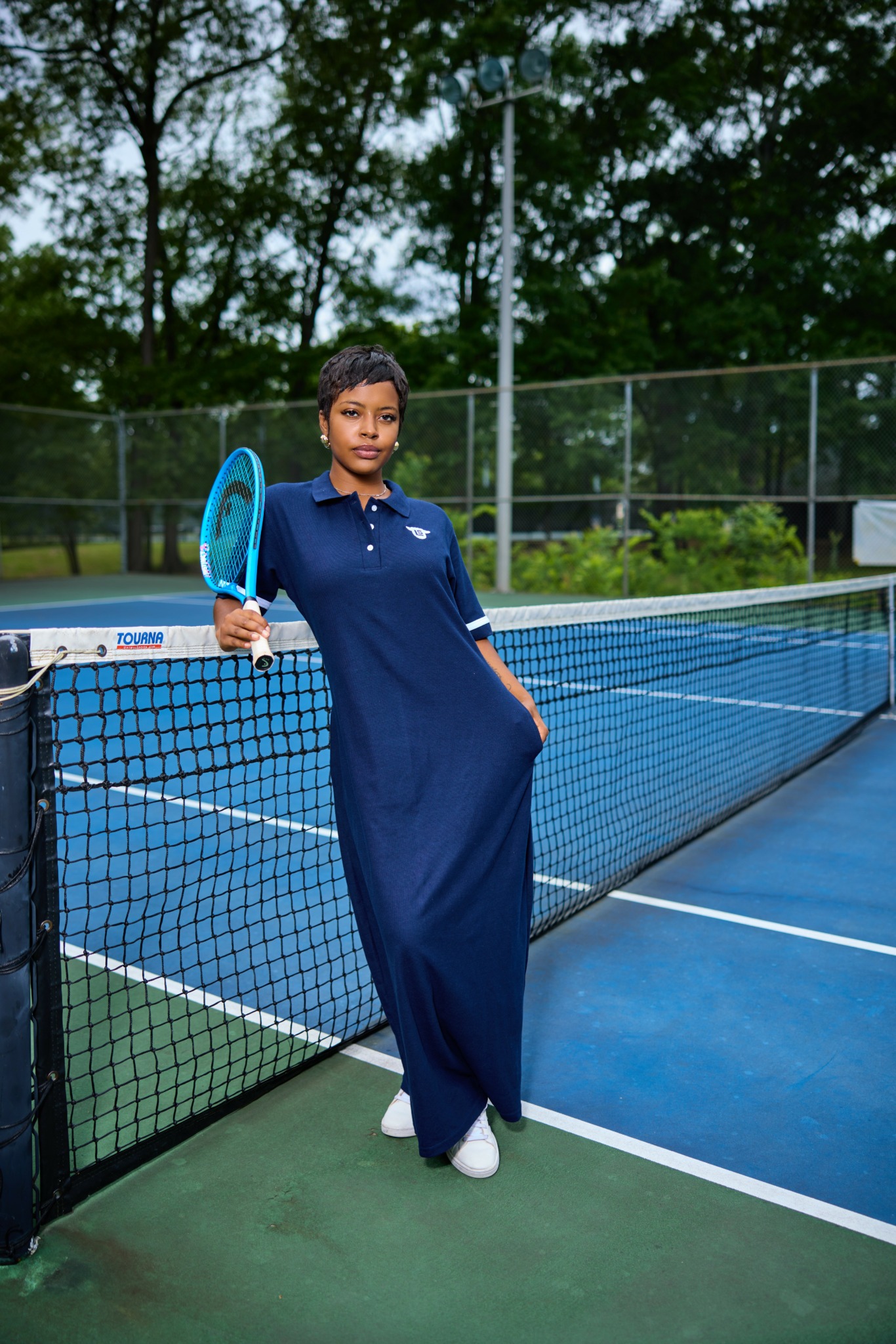 Woman in a navy dress holding a tennis racket on a tennis court, with trees and a fence in the background.
