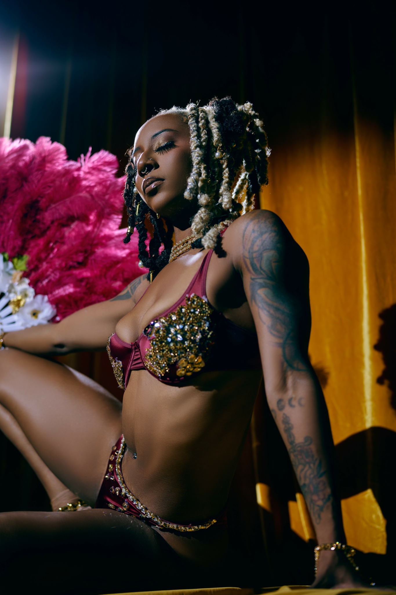 Woman with braided hair and tattoos, wearing a bejeweled top, sitting on a stage with a pink feathered prop, dark background