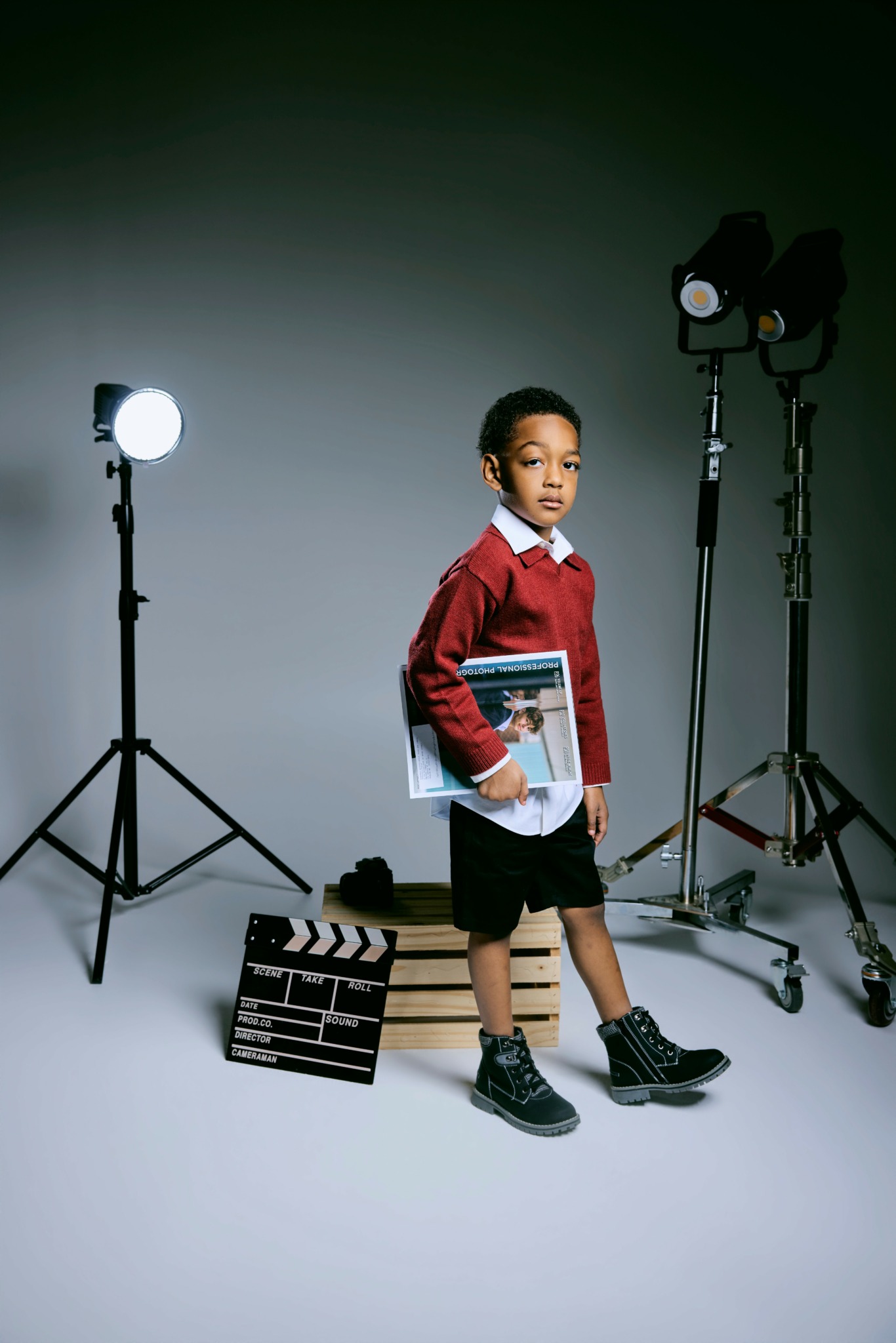Child holding a clapperboard in a photography studio with lighting equipment, standing on a plain background.