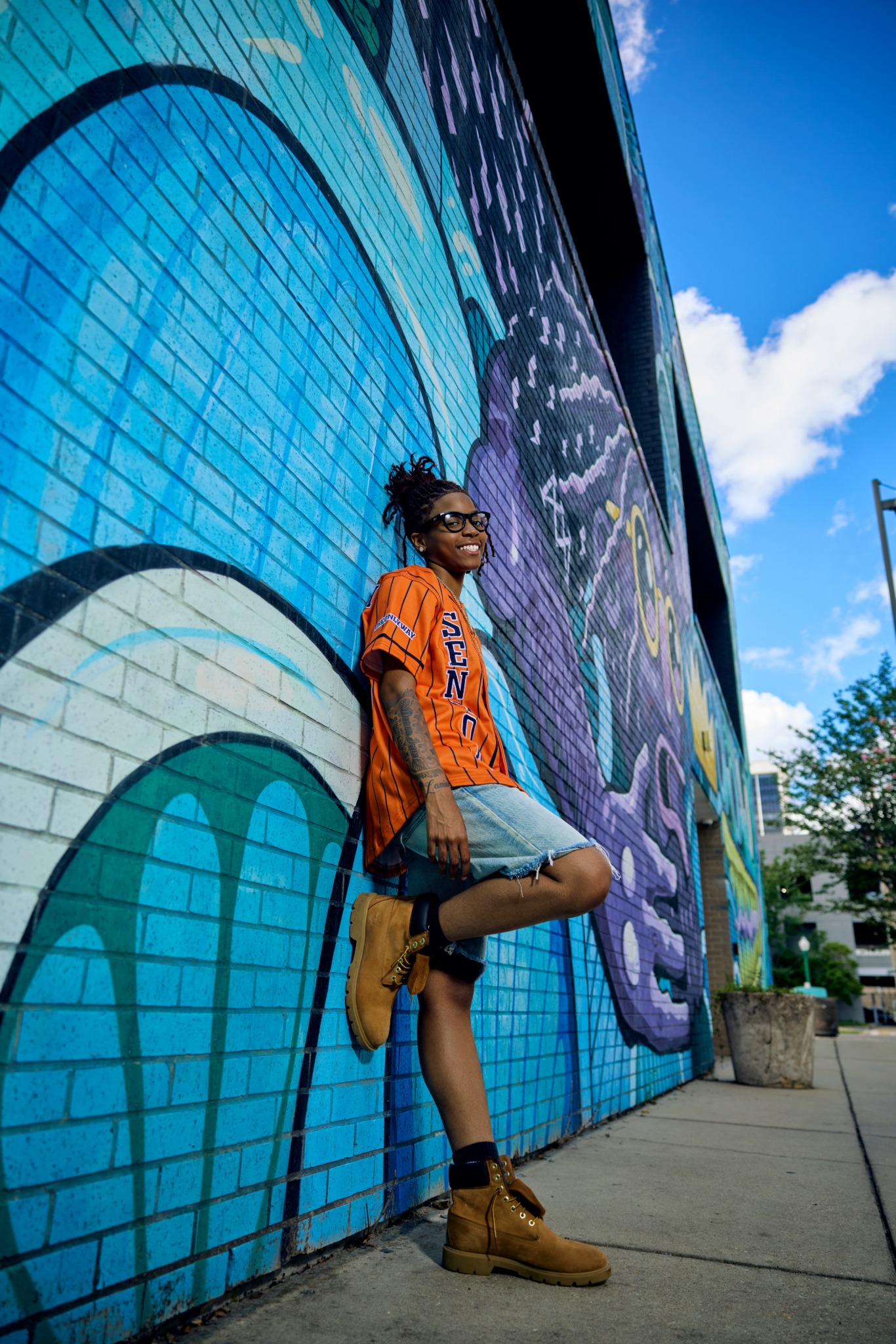 Person with glasses and orange shirt leaning against a colorful mural wall, smiling, with blue sky and clouds above.